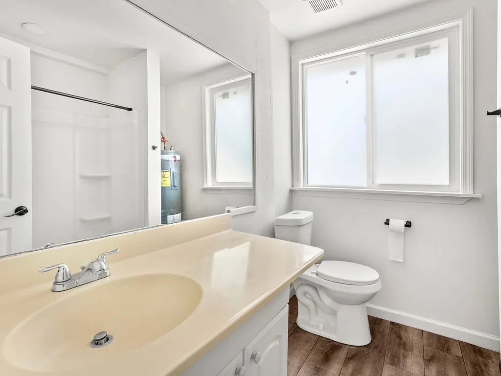 Wood-finish flooring, white vanity with integrated sink, chrome faucet, white toilet, and sliding window