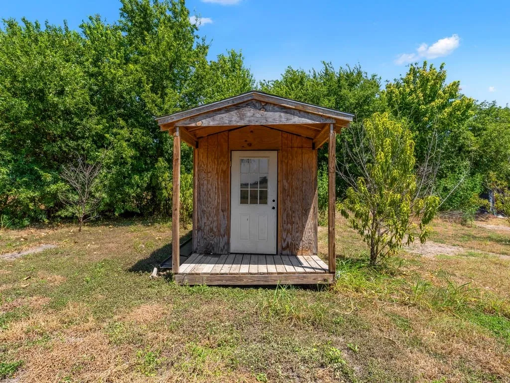 Wood-sided structure with a covered porch and white door, situated on a grassy area with mature trees in the background