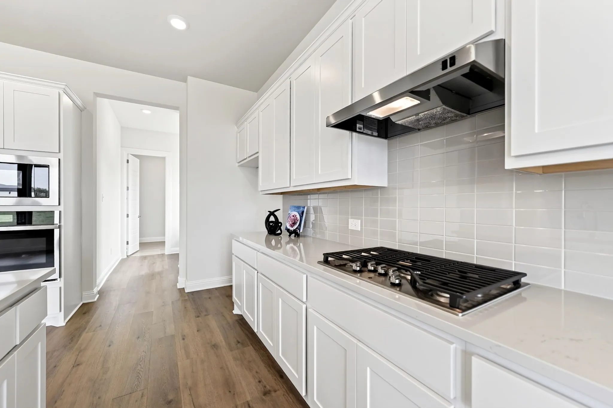 White shaker-style cabinetry, wood-finish flooring, built-in oven and microwave, integrated cooktop, and subway tile backsplash