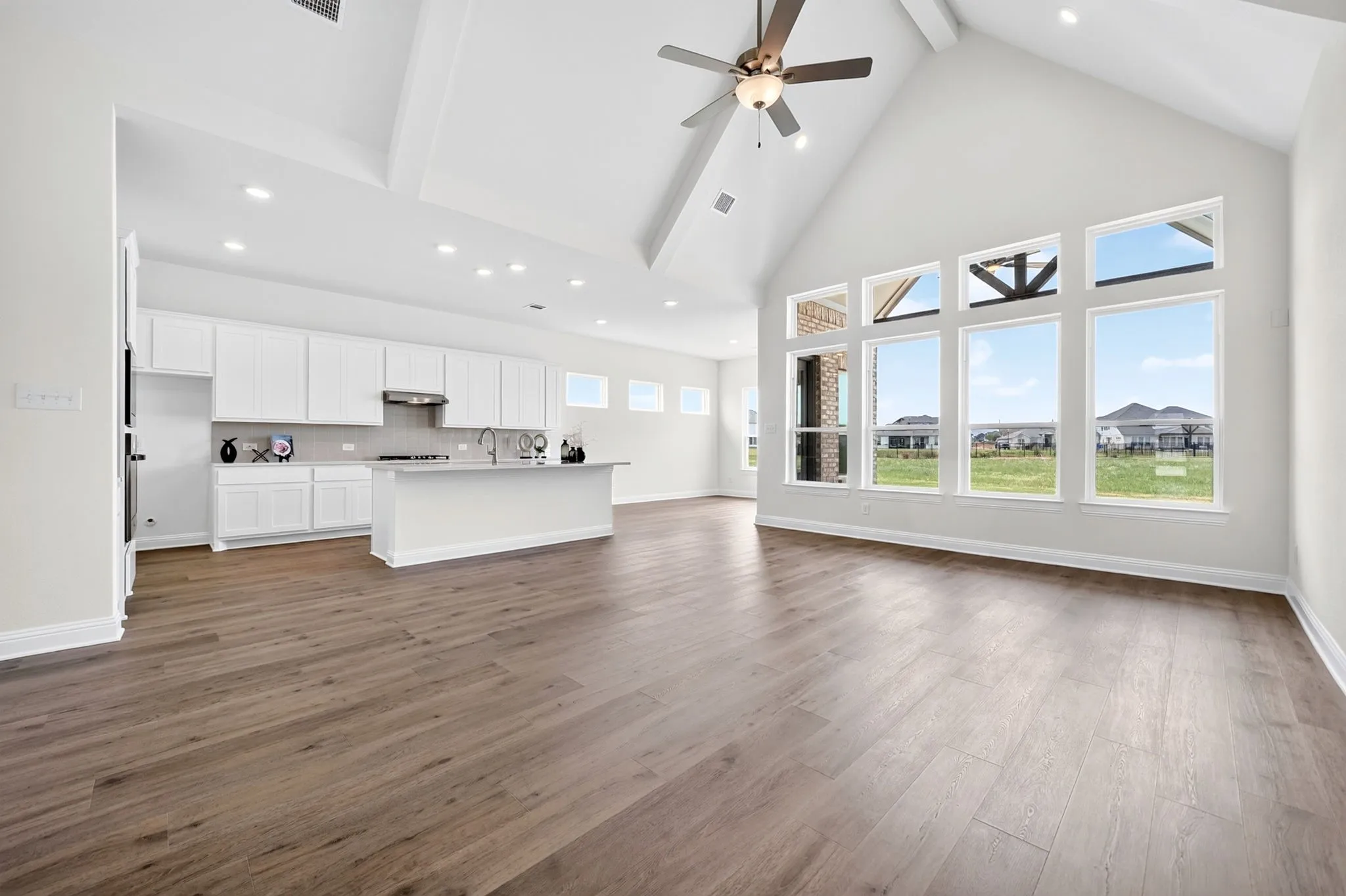 Vaulted ceiling with exposed beams, wood-finish flooring, white cabinetry, kitchen island with sink, and large windows