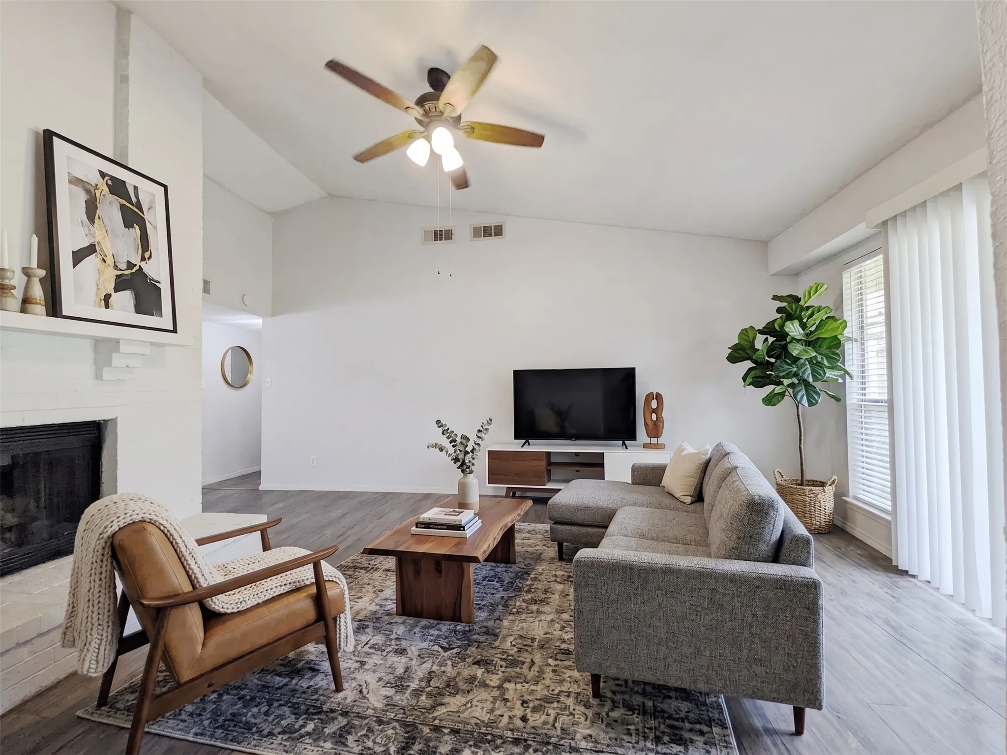 Bright, open living space featuring hardwood flooring, a wood-burning fireplace with white surround, and ceiling fan with integrated lighting