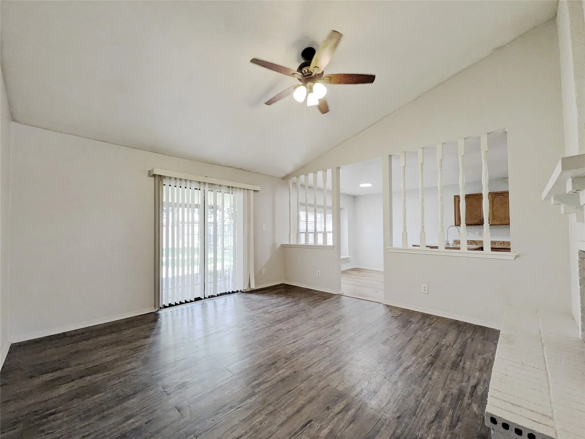Vaulted ceiling, wood-finish flooring, ceiling fan with integrated lighting, sliding glass door with vertical blinds, and a partial wall with decorative spindles