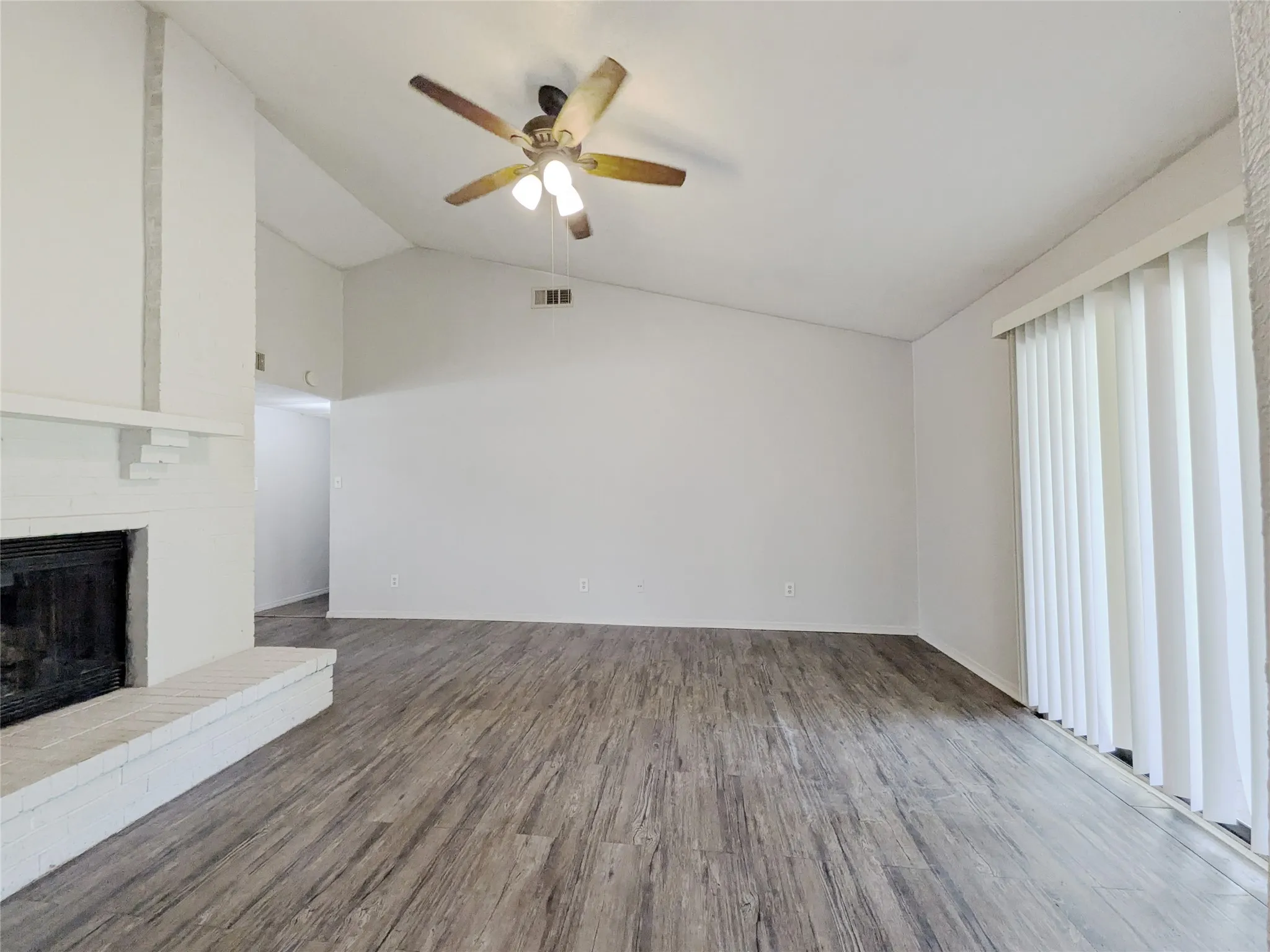 Vaulted ceiling with a ceiling fan, brick fireplace, wood-finish flooring, and vertical blinds