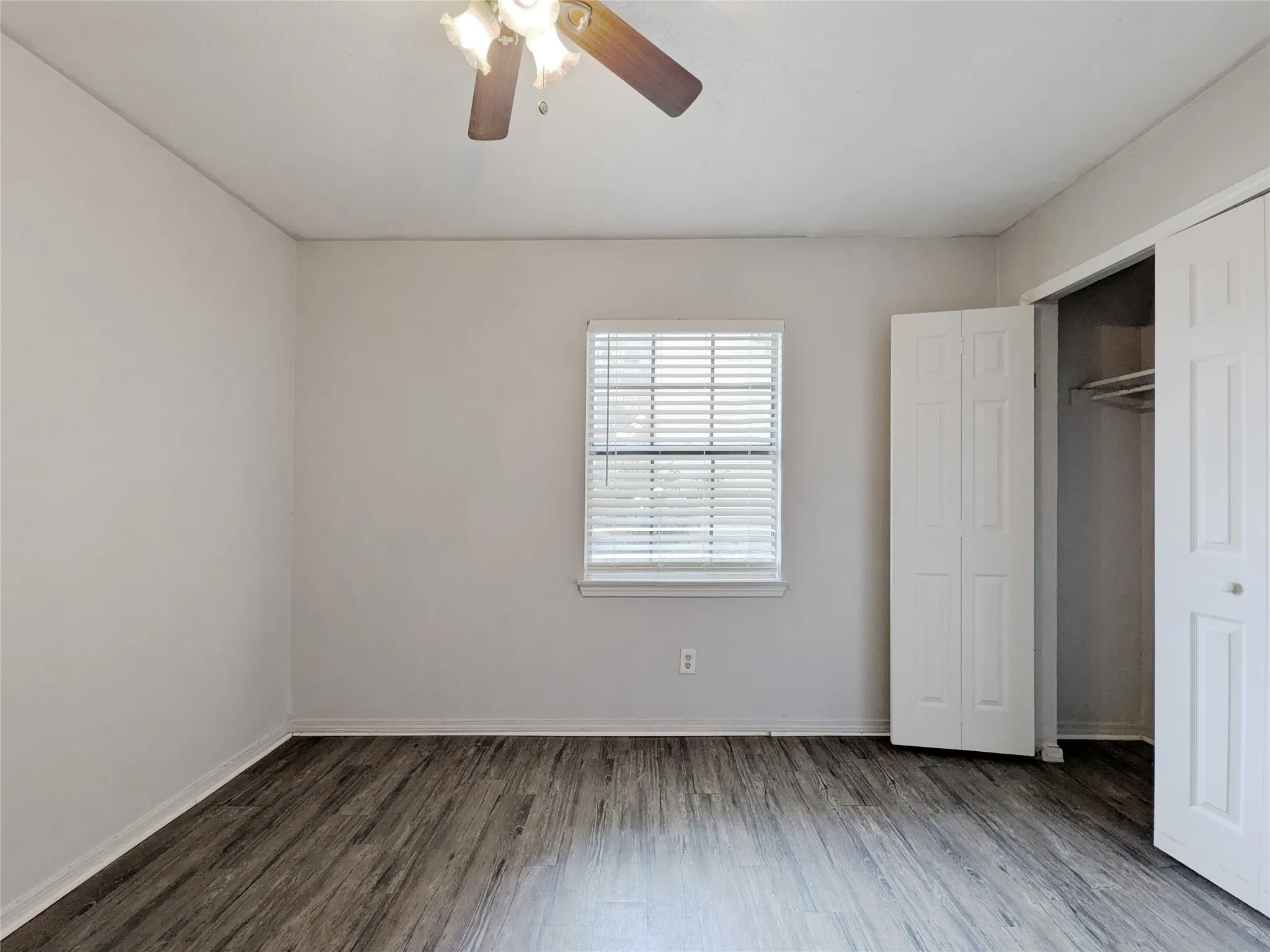 Wood-finish flooring, ceiling fan with light fixture, window with blinds, and bi-fold closet doors