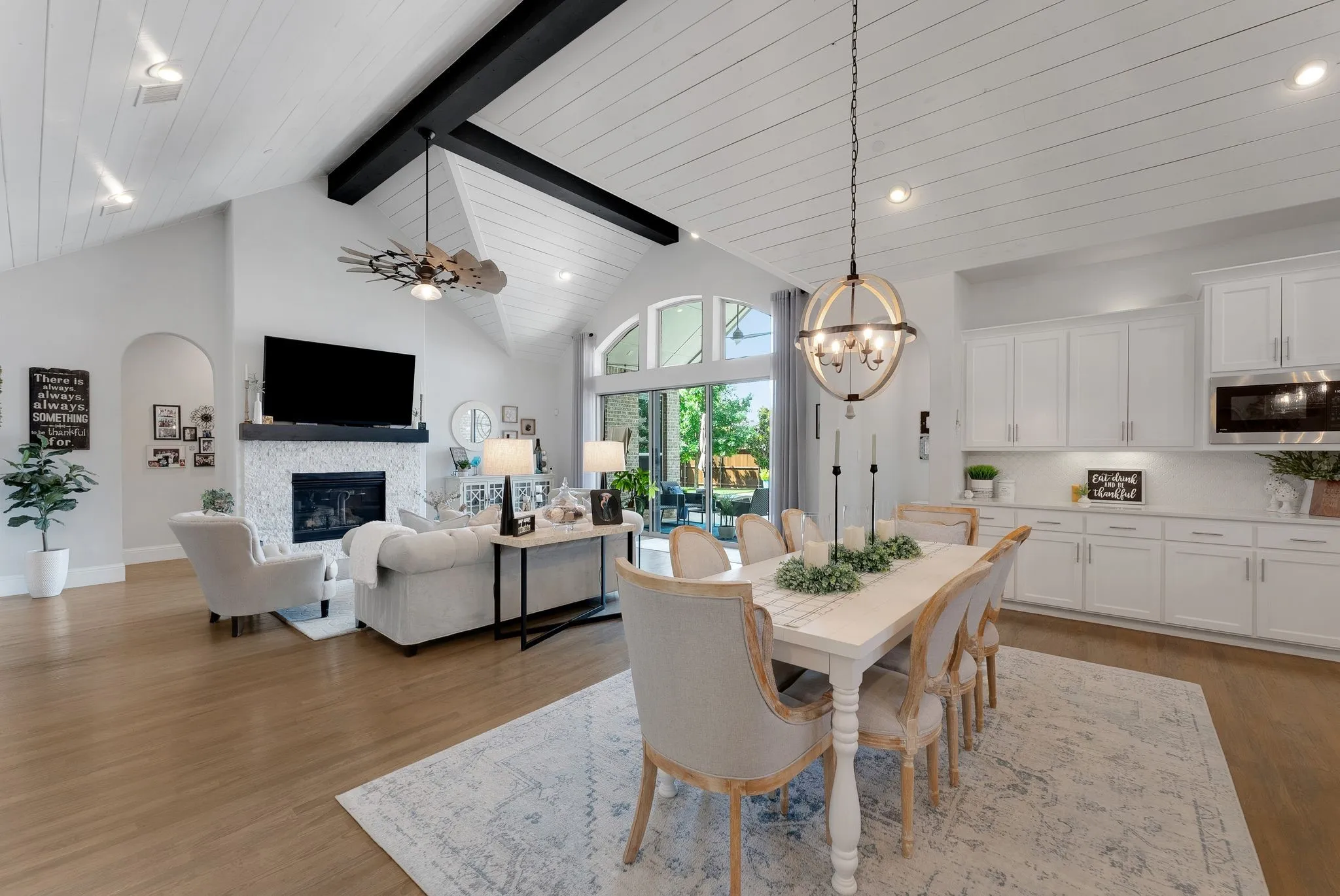 Vaulted ceilings with exposed black beams, white shiplap ceiling, fireplace with white tile surround, wood-finish flooring, and a kitchen with white cabinetry and stainless steel appliances