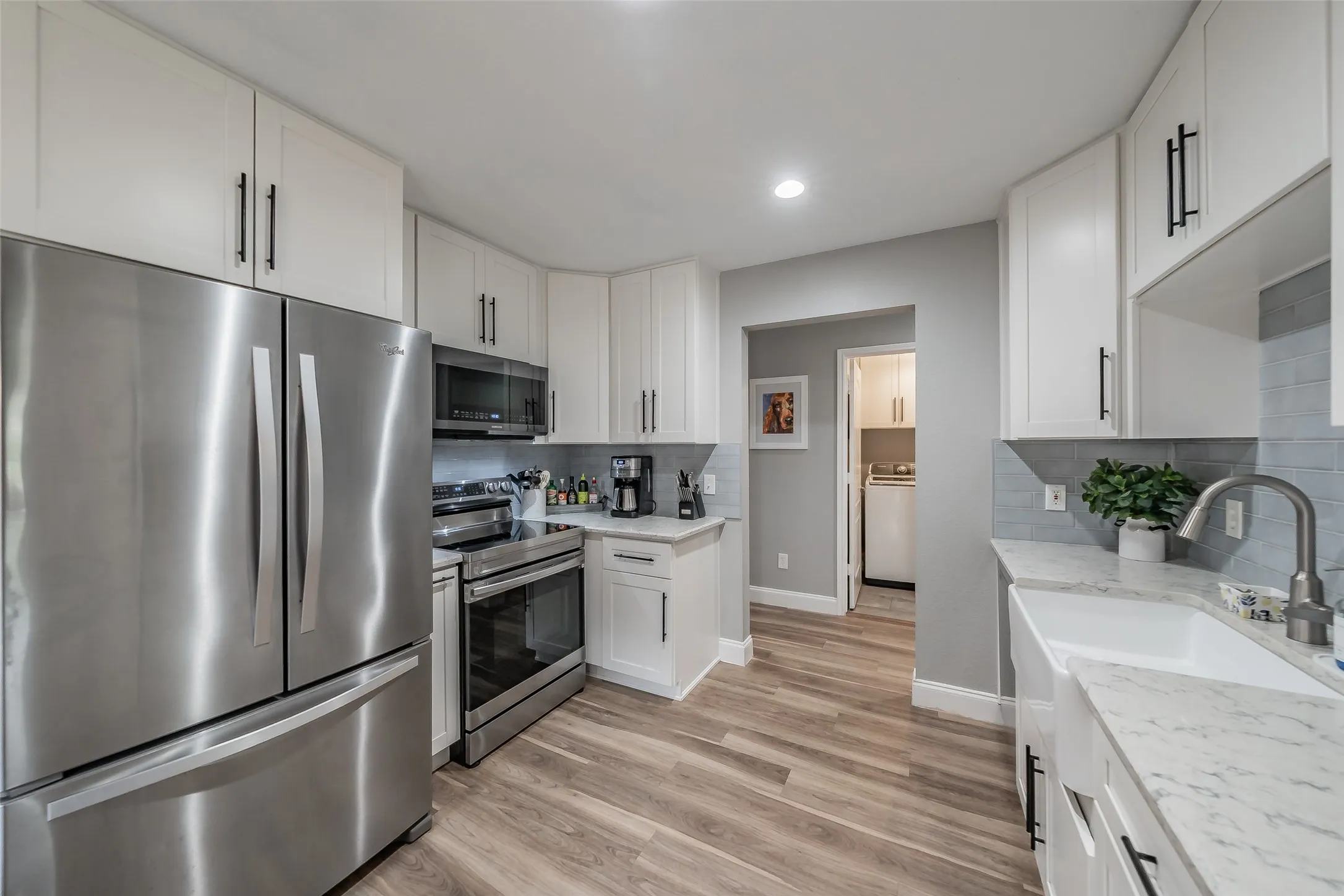 Modern kitchen featuring stainless steel appliances, white cabinetry with matte black hardware, wood-finish flooring, a farmhouse sink, and light-toned countertops