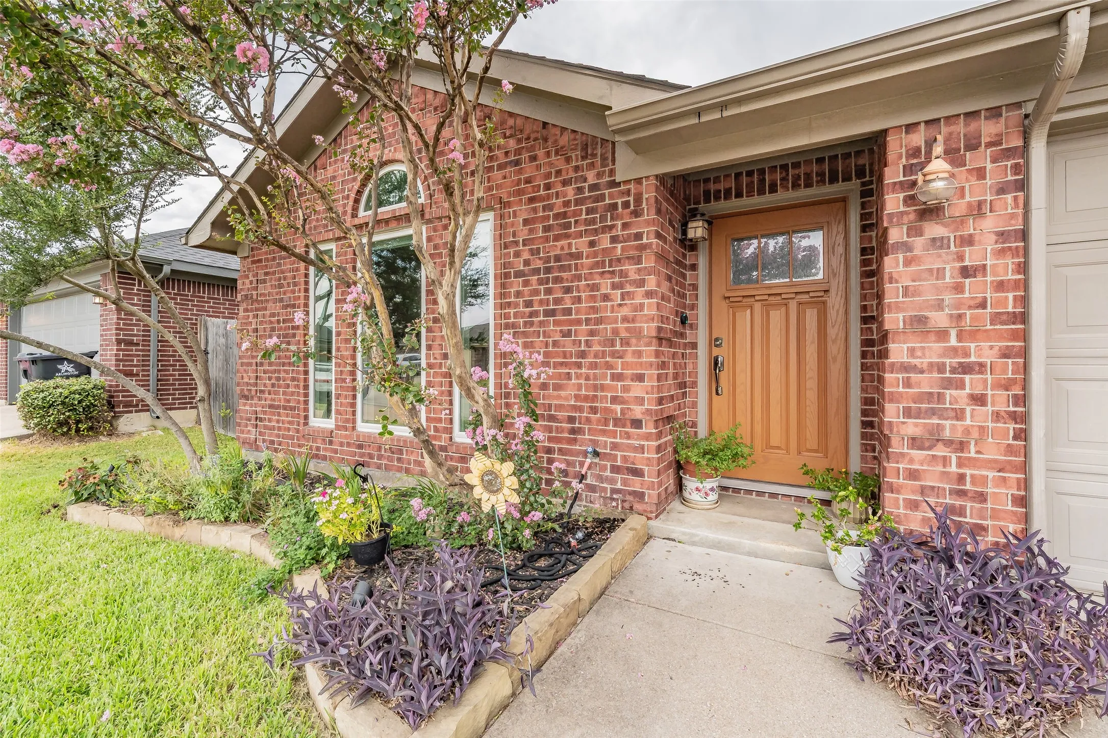 Brick exterior featuring a wood-panel entry door with transom window, arched window, and established garden beds