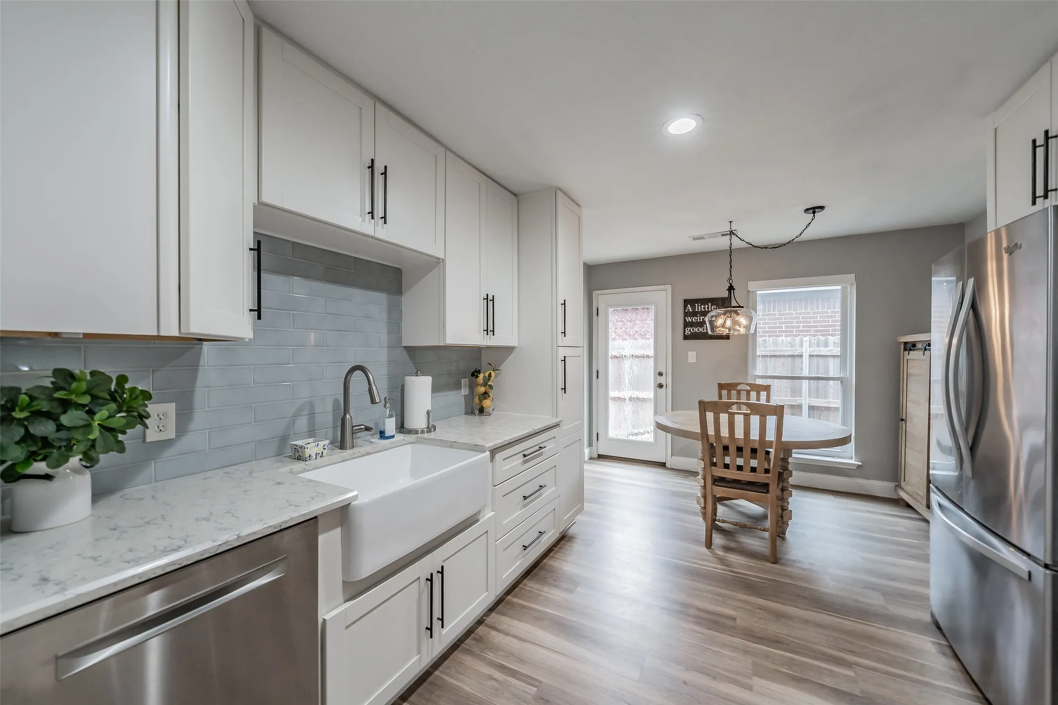 White shaker cabinetry, light-toned wood-finish flooring, stainless steel appliances, a farmhouse sink, and a tiled backsplash