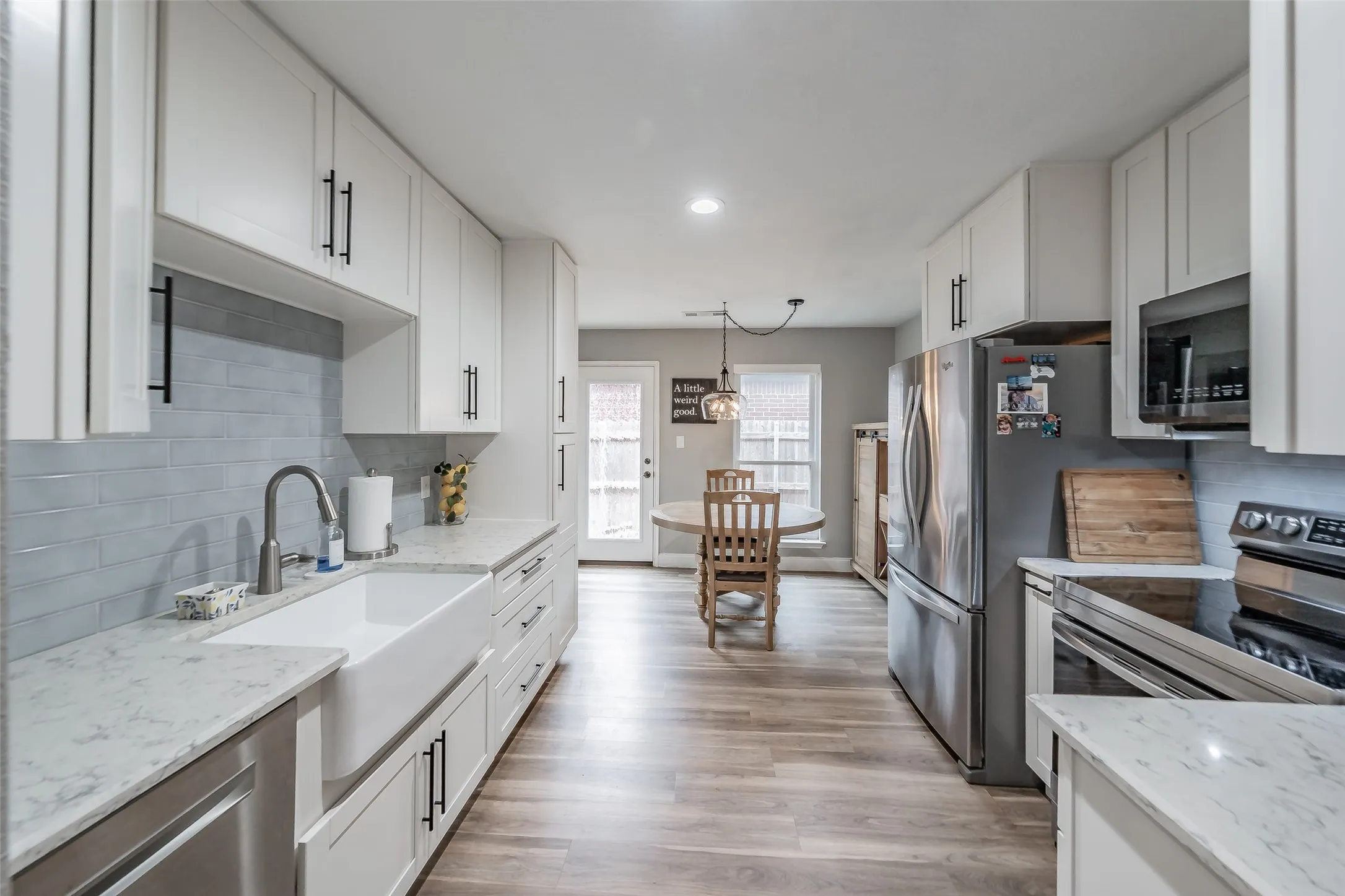 White shaker-style cabinetry with matte black hardware, light-toned wood-finish flooring, a white farmhouse sink with a gooseneck faucet, light-colored countertops, and stainless steel appliances