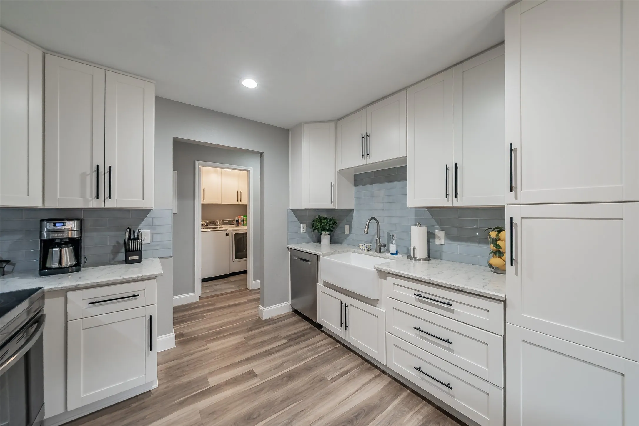 White shaker cabinetry, light-toned wood-finish flooring, white farmhouse sink, stainless steel dishwasher, and a light blue tile backsplash