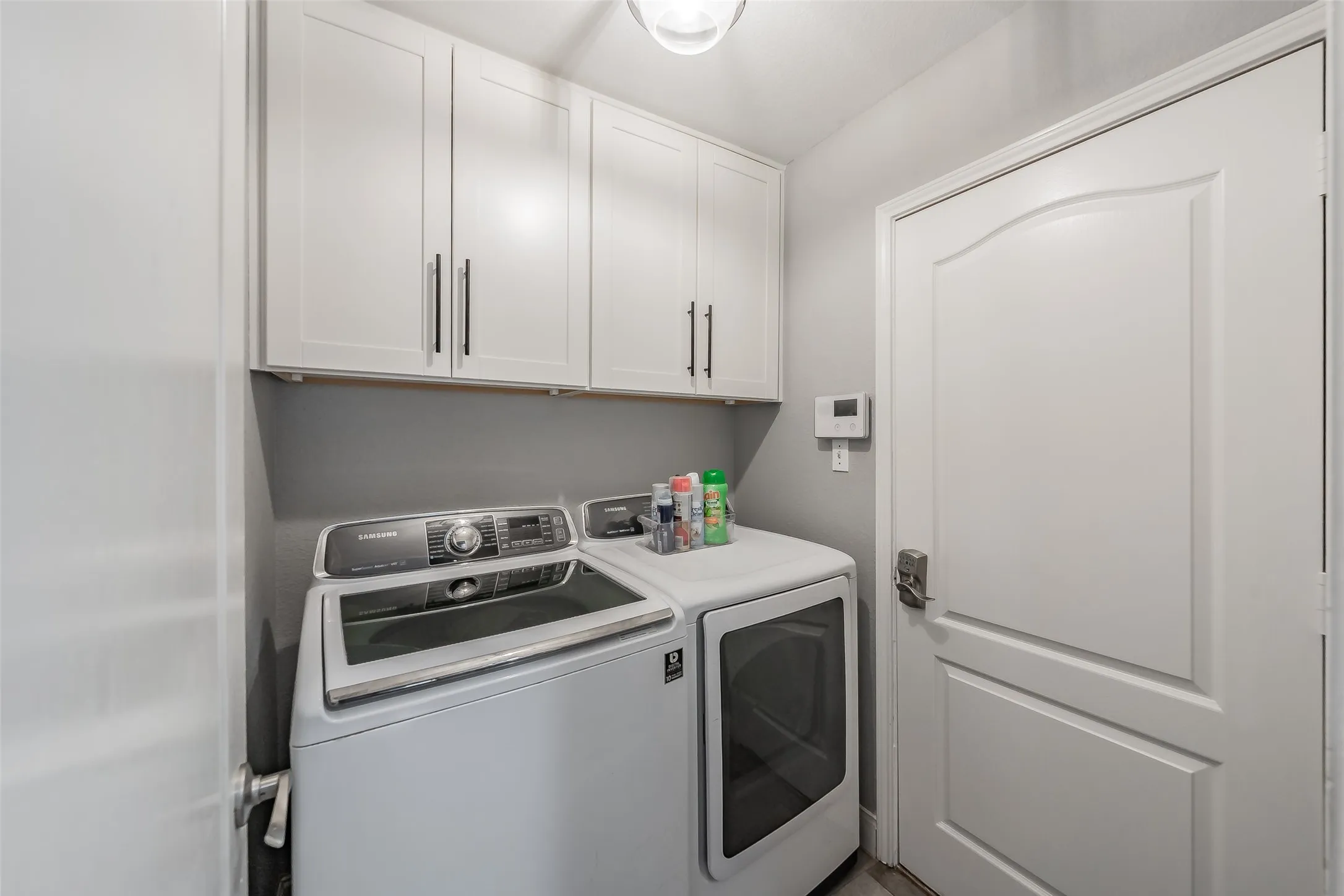 Dedicated laundry room featuring white cabinetry with stainless steel hardware, a top-load washer with digital controls, a front-load electric dryer with a large viewing window, gray tile backsplash, and white panel doors providing direct access to adjacent spaces
