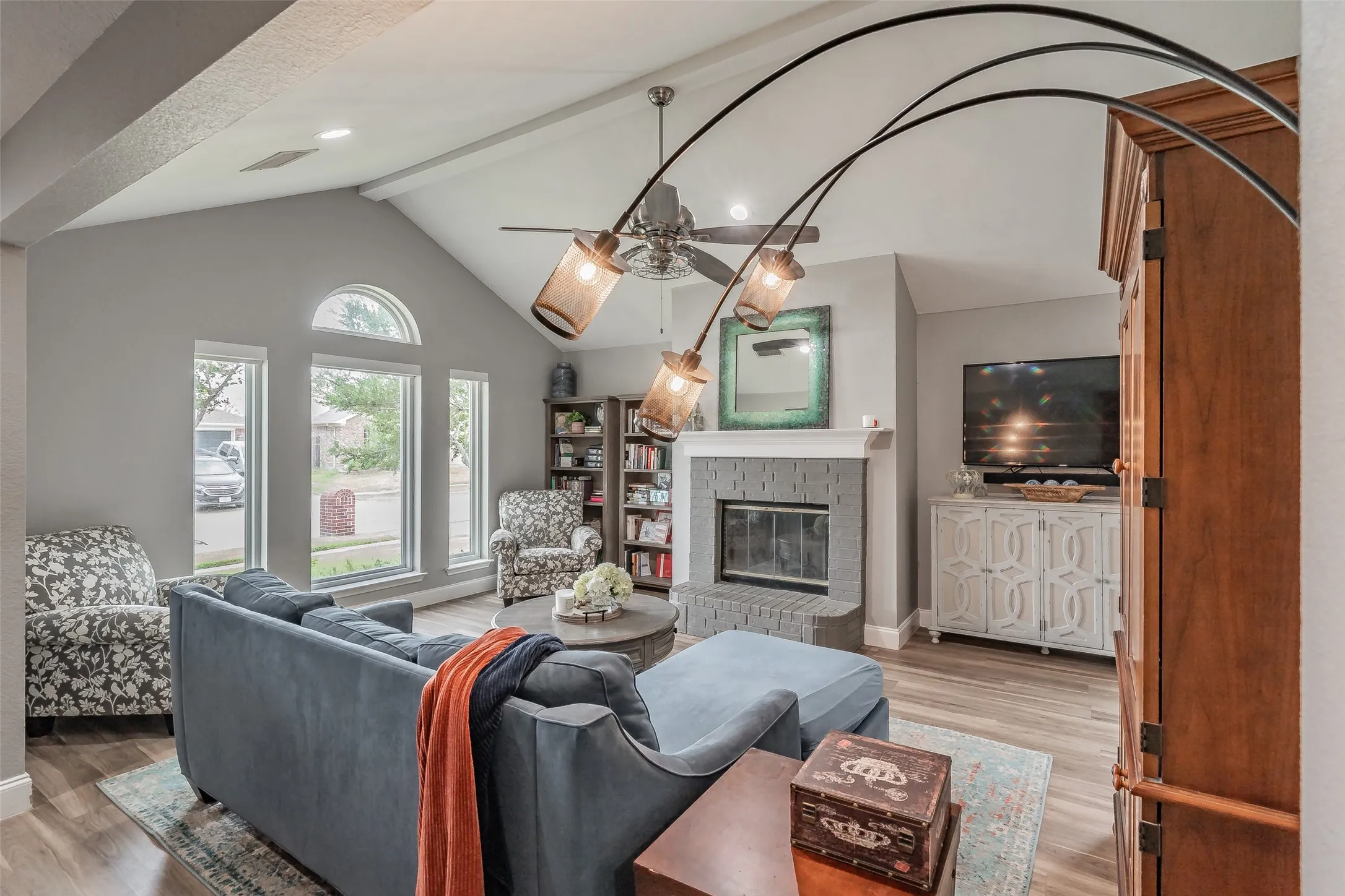 Vaulted ceiling, ceiling fan, wood-finish flooring, fireplace with gray brick surround, and multiple windows