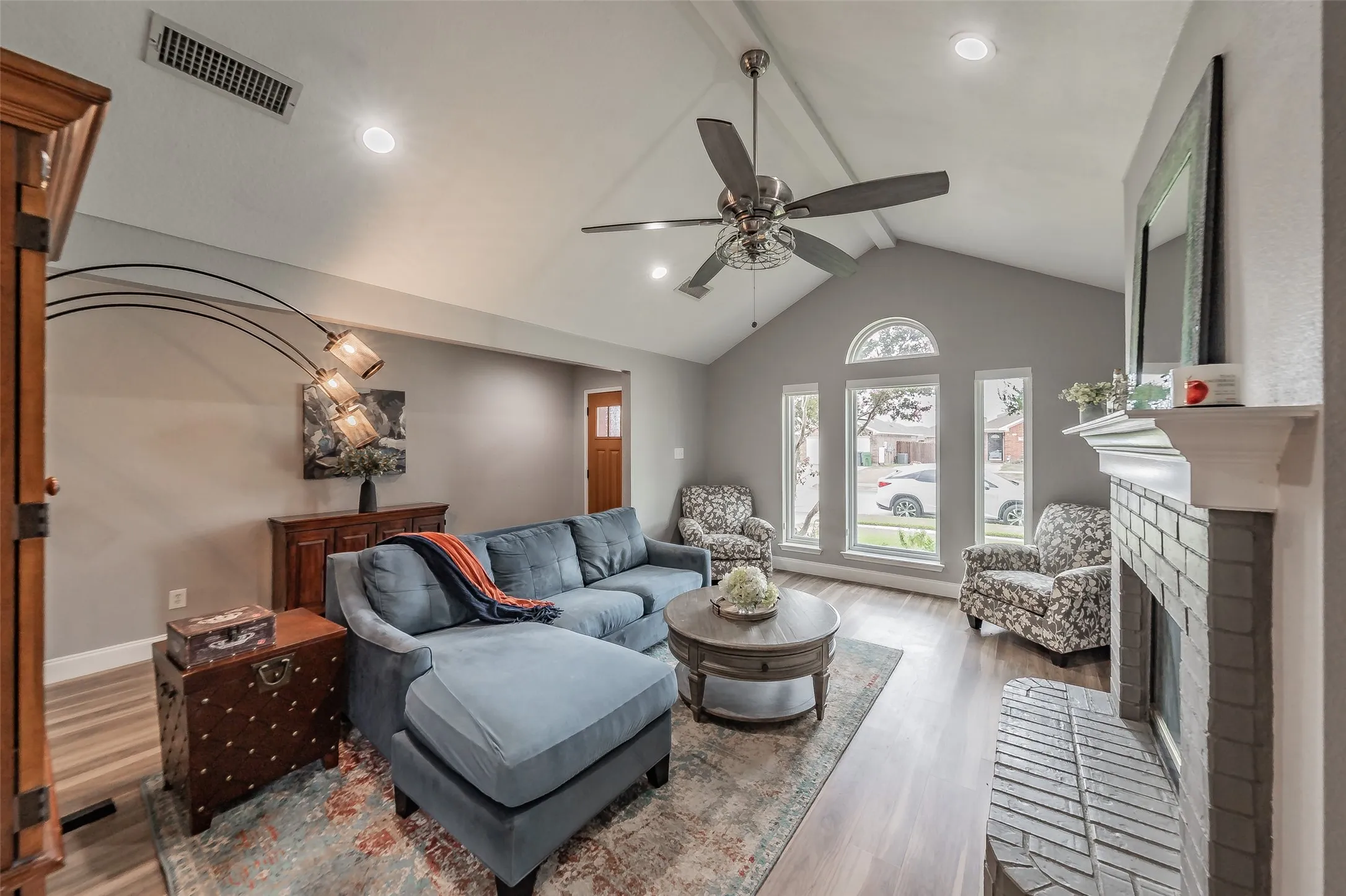 Vaulted ceiling with recessed lighting, large arched window, wood-finish flooring, brick fireplace surround, and ceiling fan