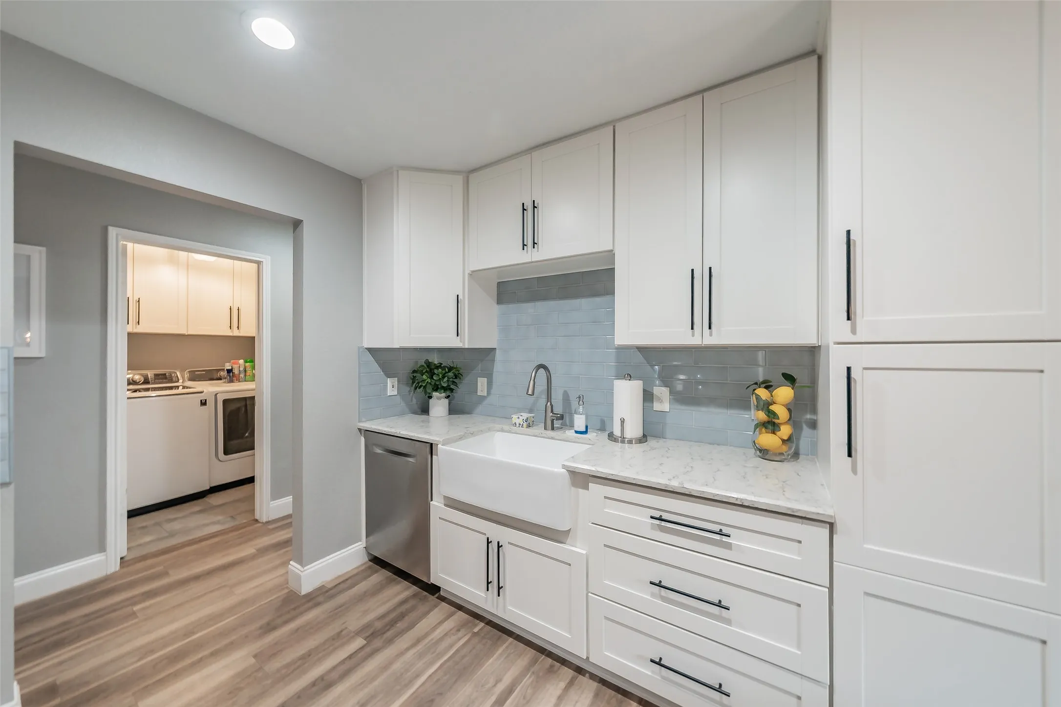 White shaker cabinetry, integrated stainless steel dishwasher, farmhouse sink, light-toned wood-finish flooring, and a blue tile backsplash