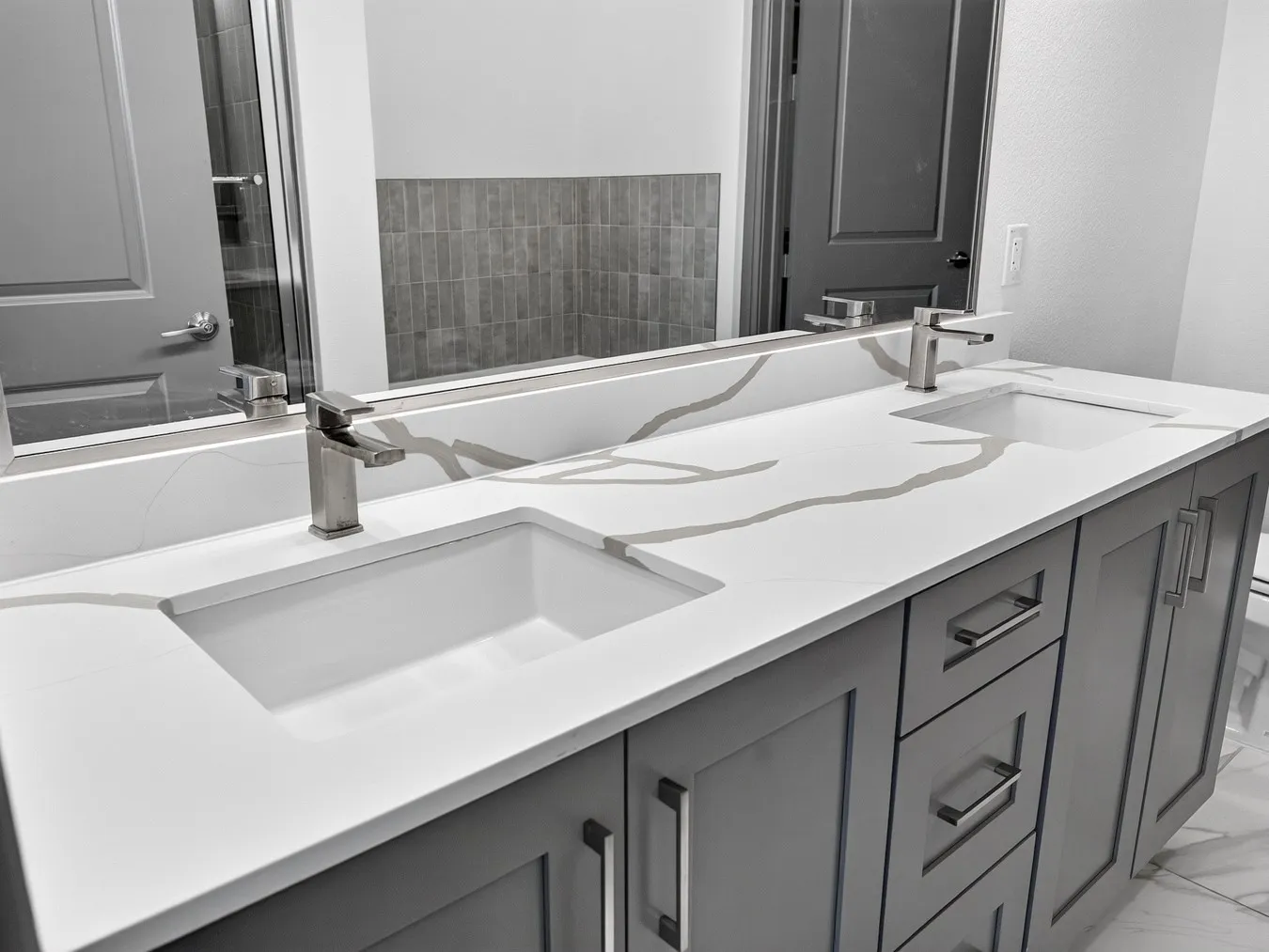 Dual undermount sinks, quartz countertop with veining, grey shaker-style cabinetry, brushed nickel hardware, and a large wall-mounted mirror