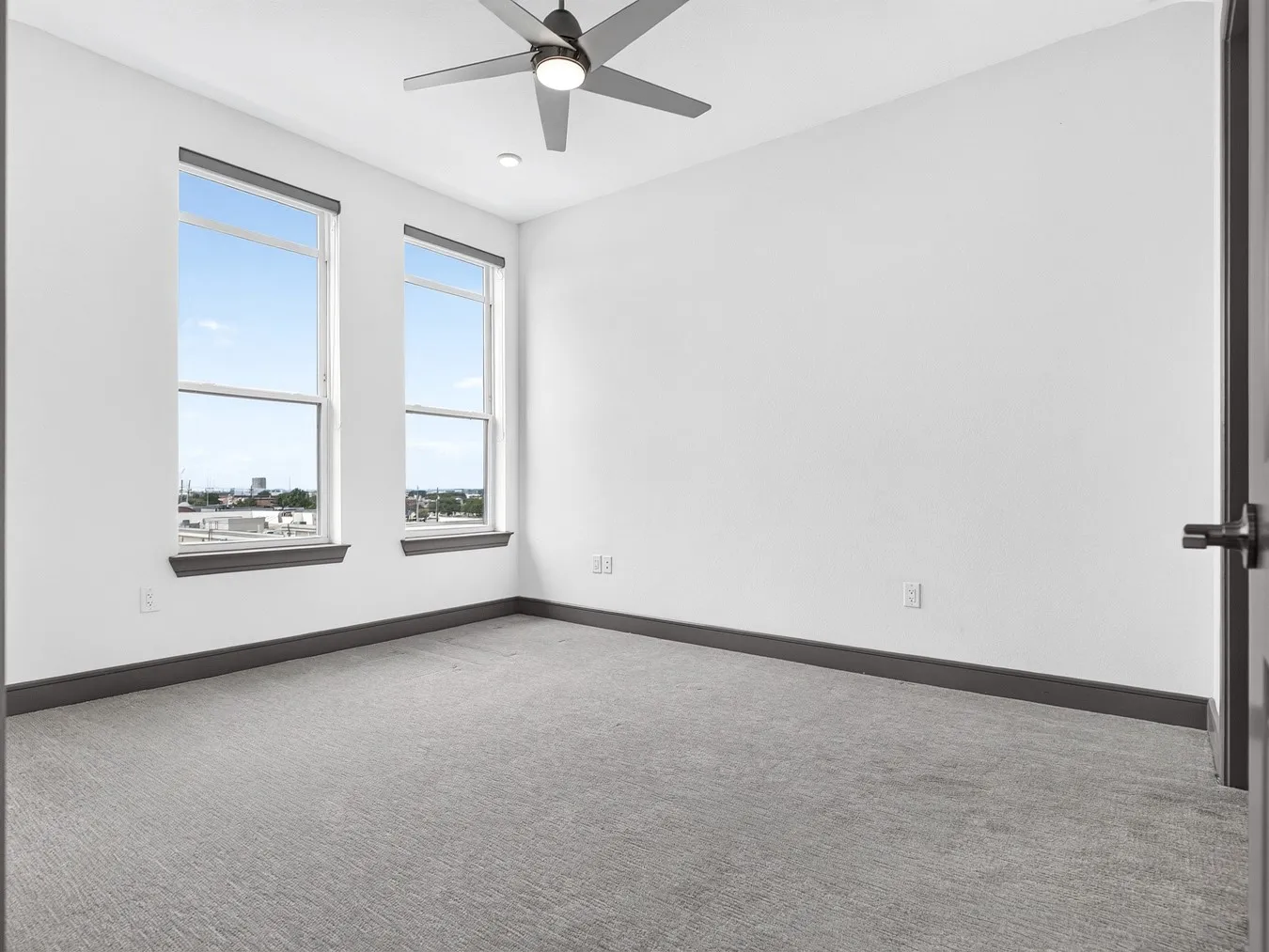 Contemporary ceiling fan with integrated lighting, two large windows, gray wall-to-wall carpeting, and dark trim