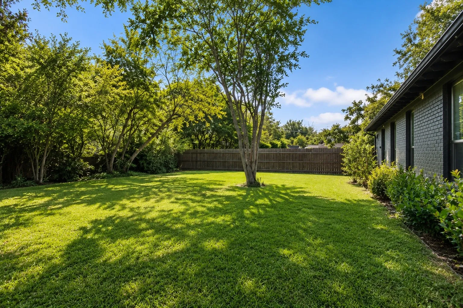 Expansive lawn with mature trees and a privacy fence, complemented by a brick exterior and garden beds