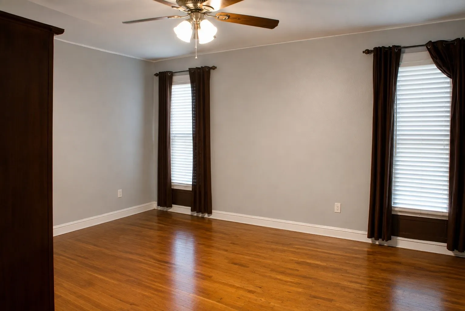 Hardwood flooring throughout, neutral wall paint, two windows with blinds, and a ceiling fan with integrated lighting
