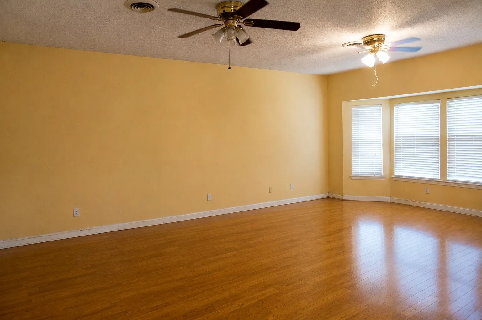 Wood-finish flooring with baseboards, bay window with blinds, and two ceiling fans with integrated lighting