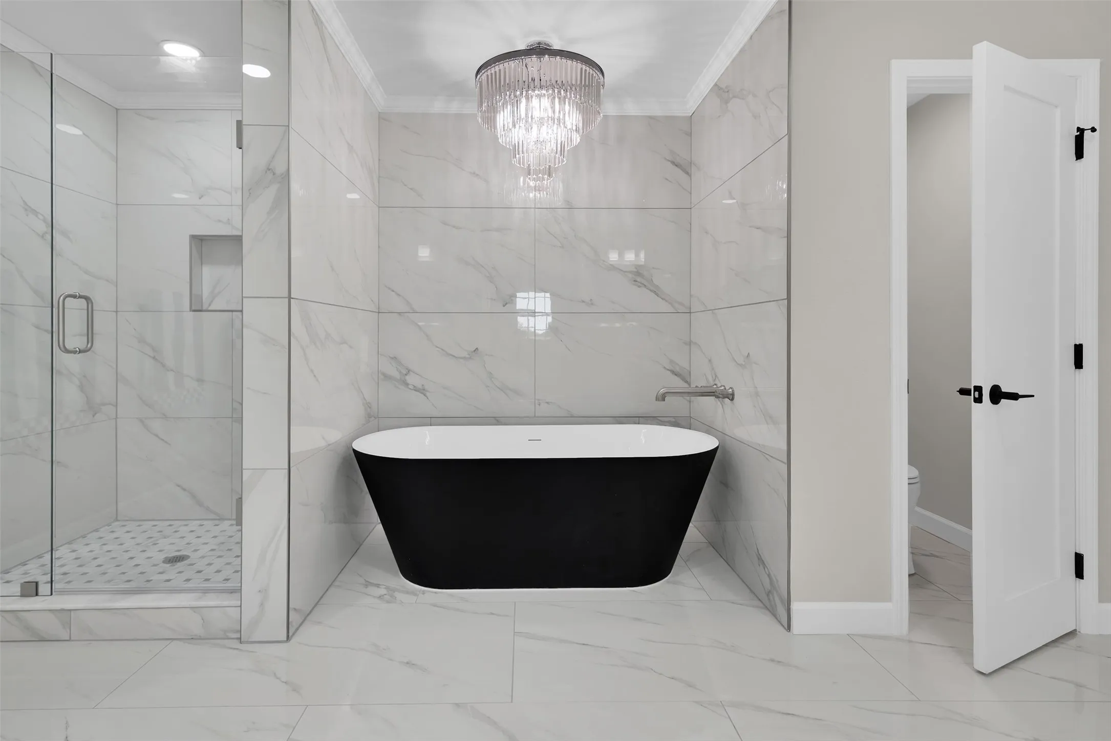 Freestanding black soaking tub, glass-enclosed shower with tiled walls and niche, large format tile flooring, and a tiered chandelier