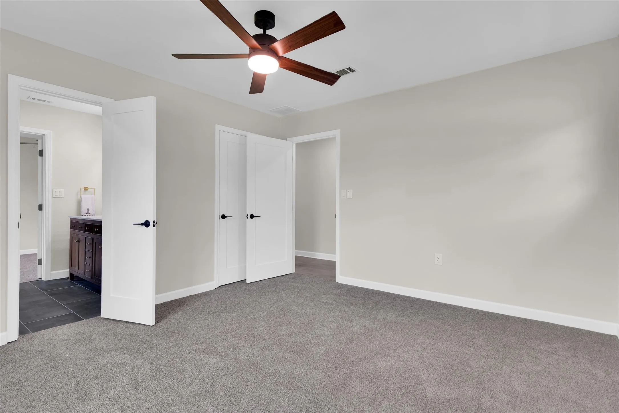 Recessed lighting and a ceiling fan with wood-finish blades illuminate a room with gray carpeting and light-toned walls