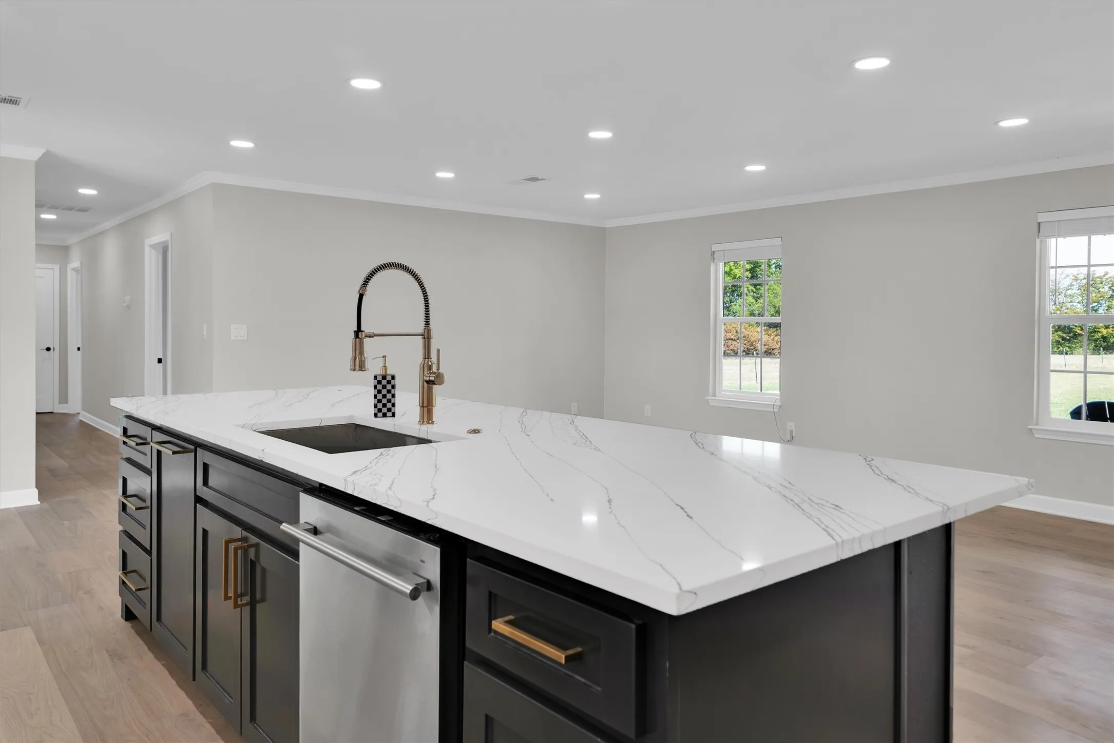 Kitchen island with a white veined countertop, integrated sink with a gold-tone pull-down faucet, stainless steel dishwasher, and dark cabinetry with gold-tone hardware