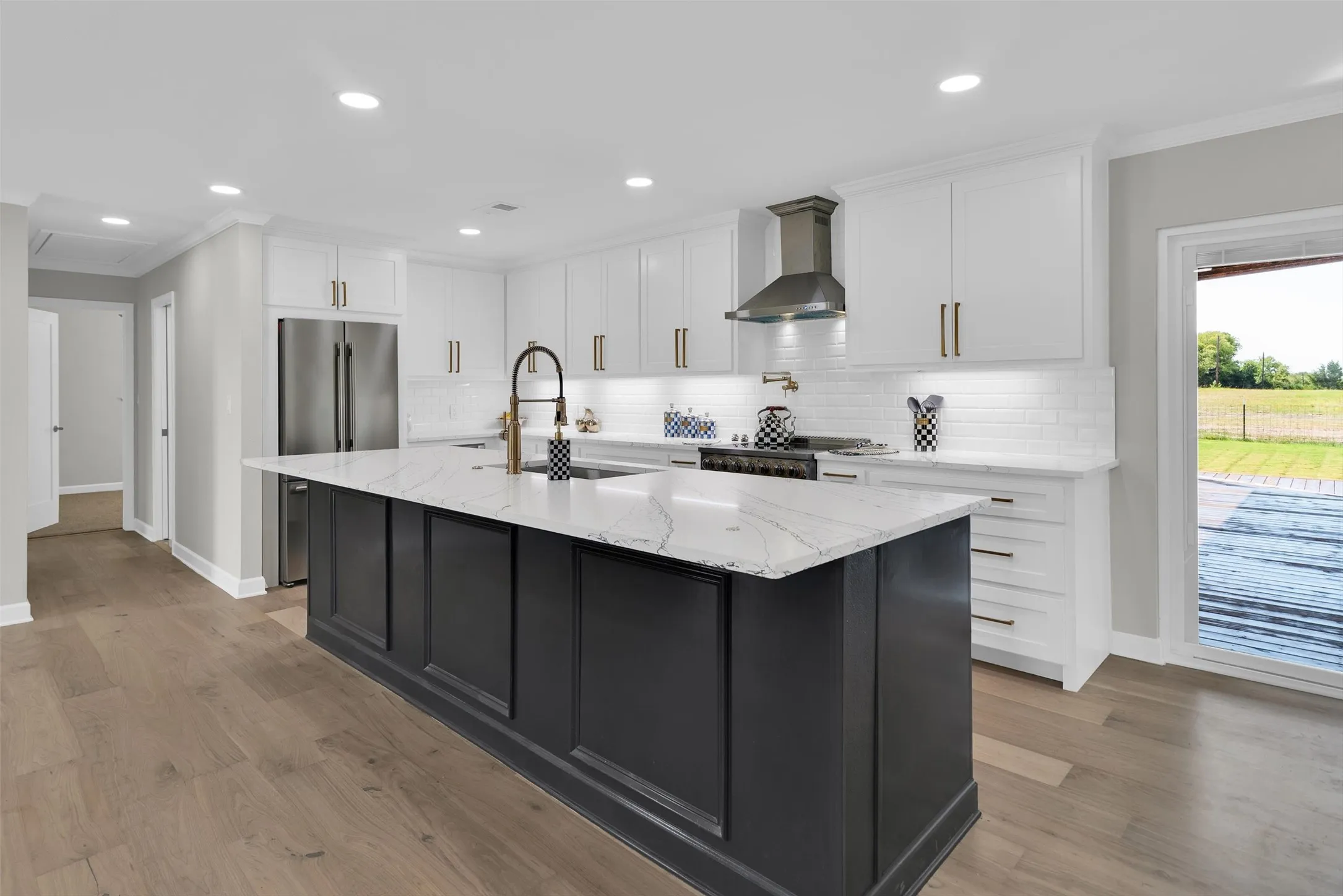 Contemporary kitchen featuring a large island with a light-toned countertop, white cabinetry with gold-toned hardware, stainless steel appliances, and wood-finish flooring