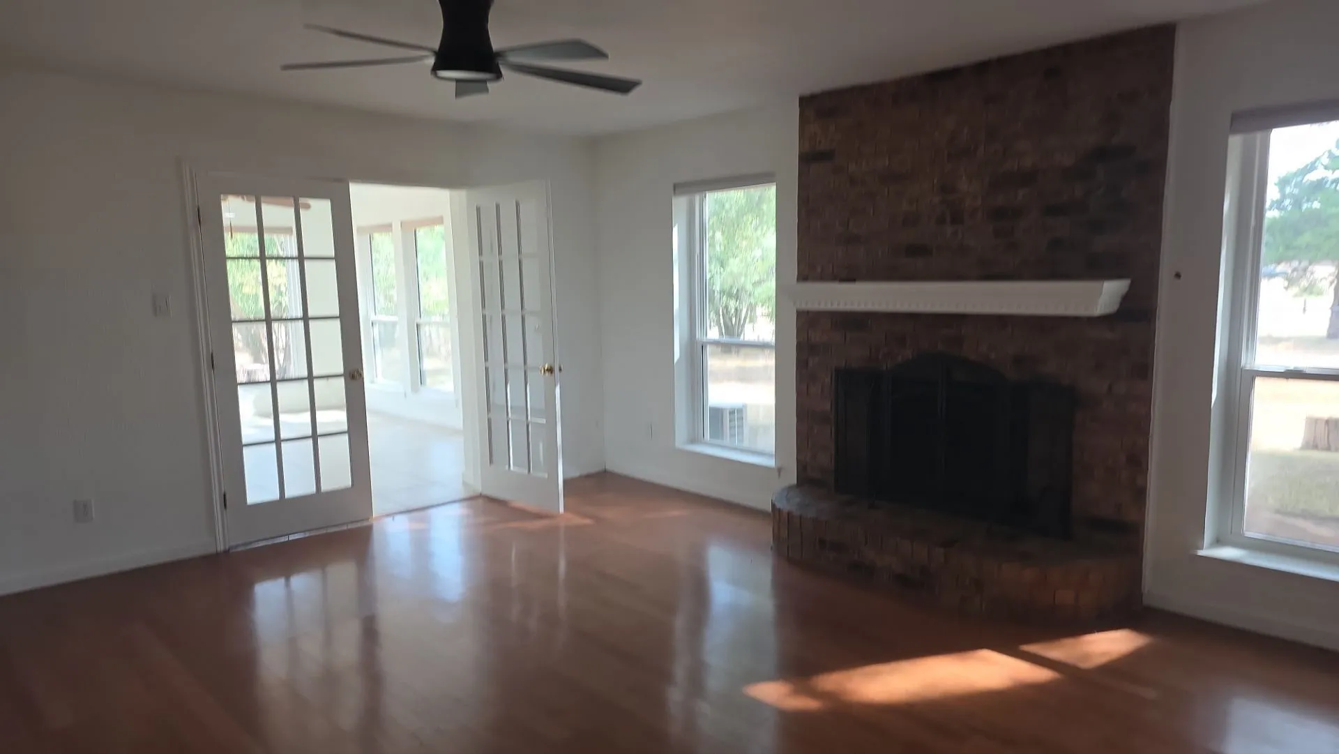 Hardwood flooring, brick fireplace with mantel, ceiling fan, and French doors opening to an additional room