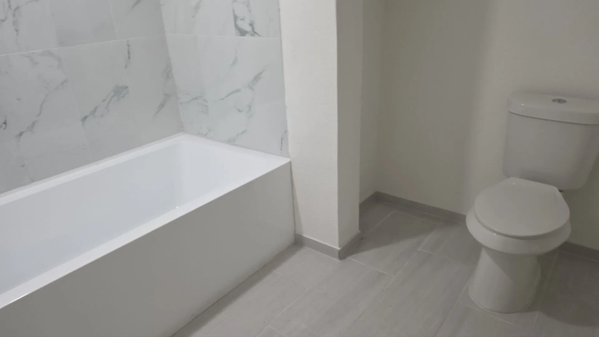 White soaking tub with marble-patterned wall tile and light-toned tile flooring