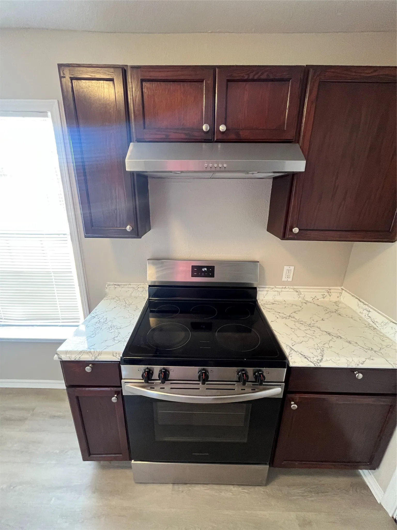Stainless steel electric range with cooktop, stainless steel range hood, dark wood-finish cabinetry, and white-patterned countertops