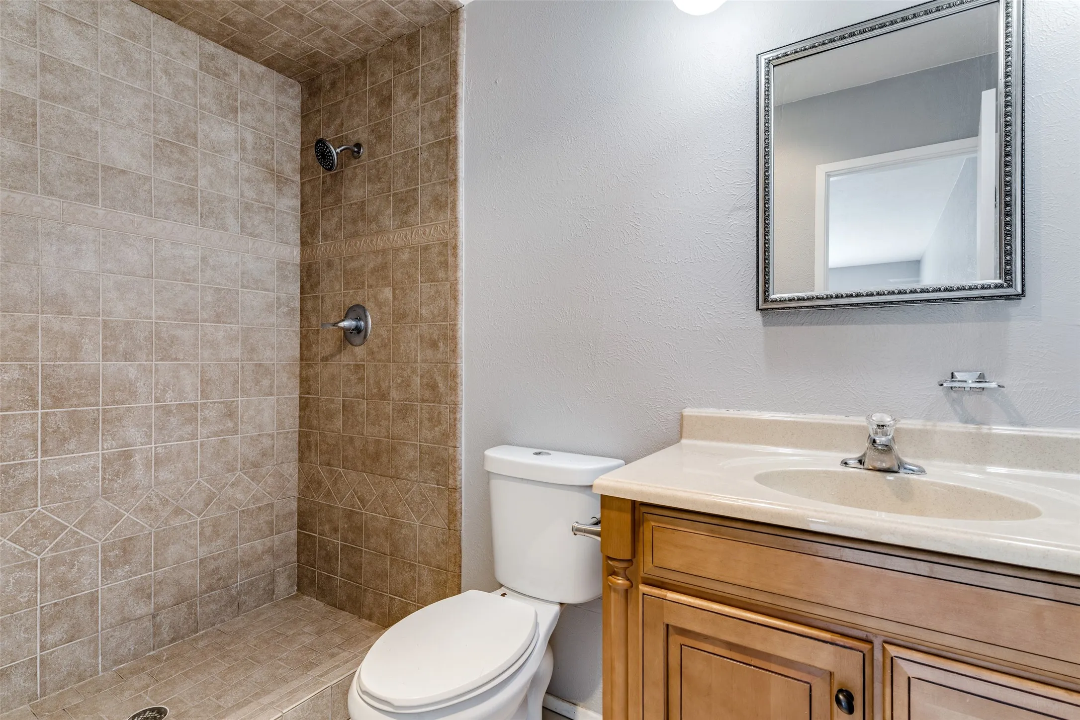 Full bathroom featuring a tiled walk-in shower with chrome fixtures, a wood-tone vanity with cultured marble countertop and integrated sink, framed mirror and toilet