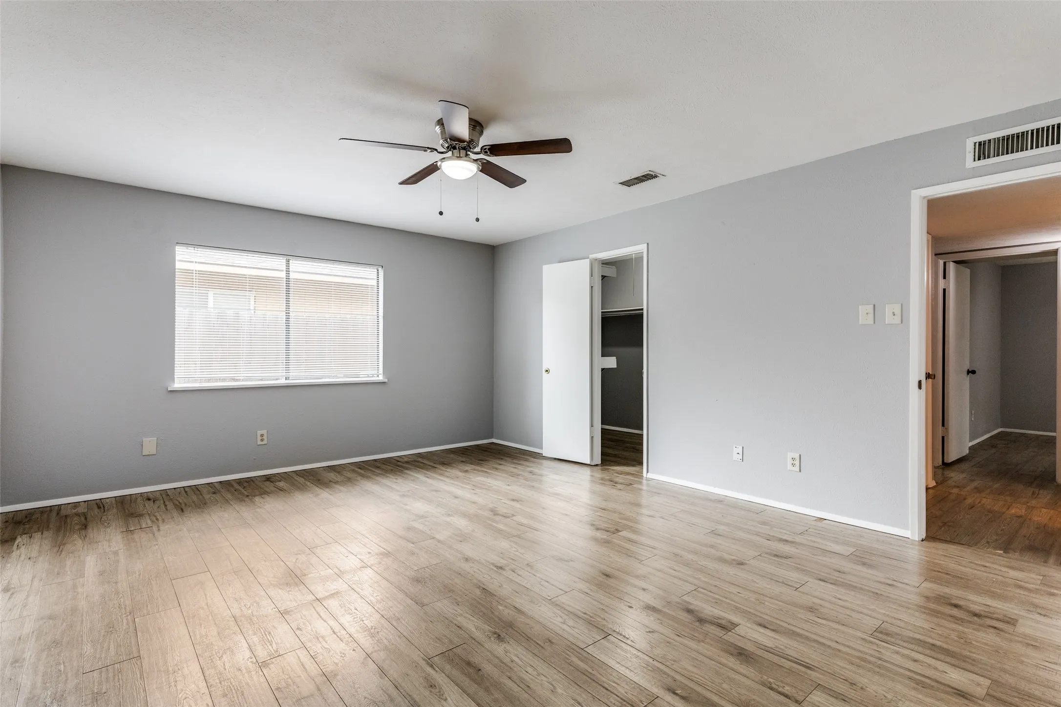 Spacious room featuring wood-finish flooring, a ceiling fan with integrated lighting, and a large window with blinds