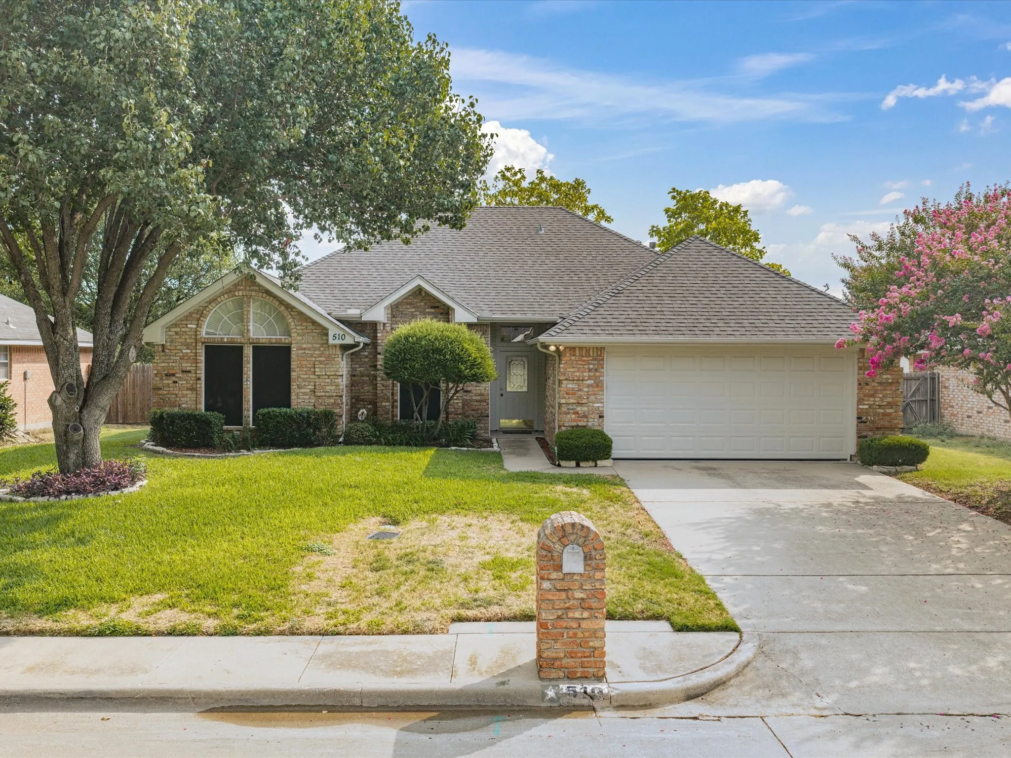 Brick exterior with an arched window, attached two-car garage, concrete driveway, and mature landscaping