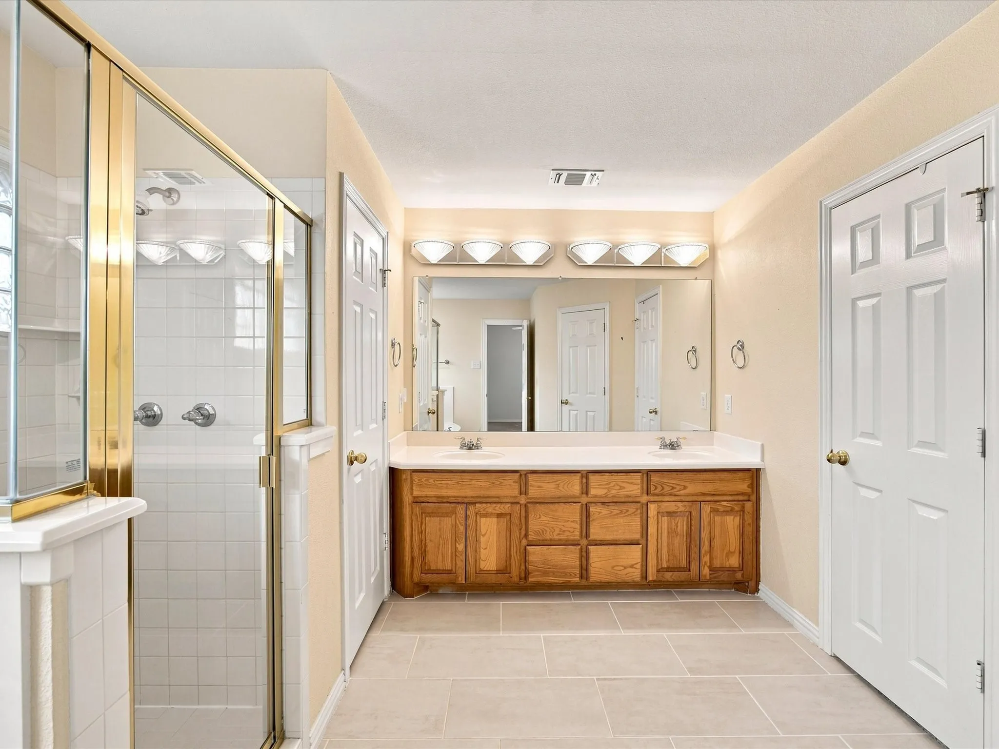 Double vanity with wood-finish cabinetry, a large integrated mirror, and a tile shower with a gold-tone framed glass enclosure