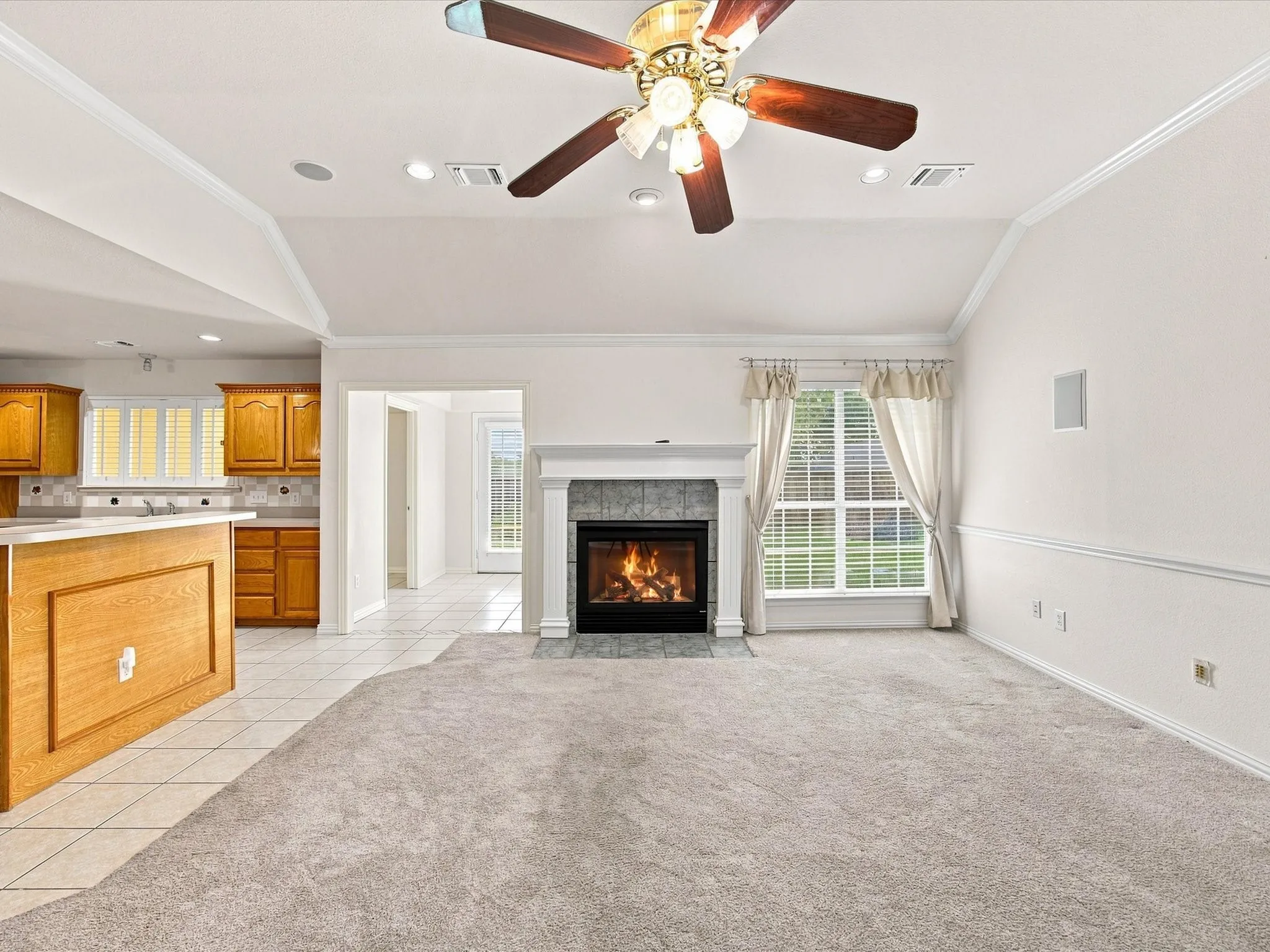 Vaulted ceiling with crown molding, ceiling fan, carpet flooring, a fireplace with a tile surround, and a kitchen with wood cabinetry