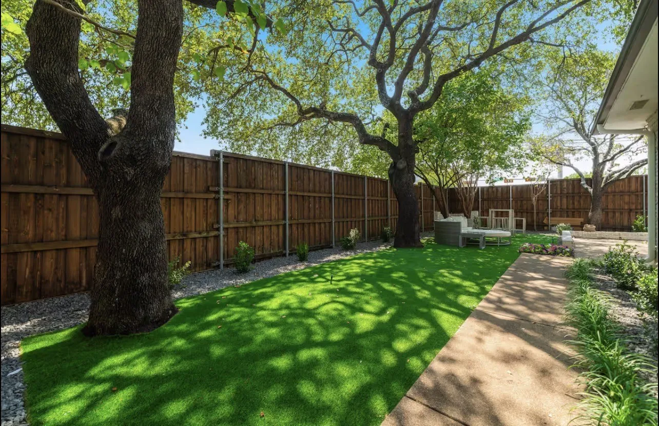Expansive artificial turf lawn with mature trees, wood fence perimeter, and gravel landscaping
