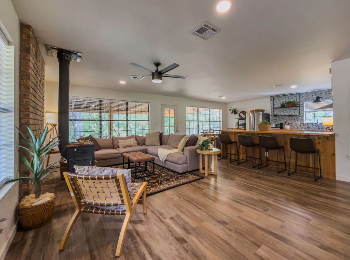 Wood-finish flooring throughout, prominent brick accent wall, wood-burning stove, large windows, and a breakfast bar with wood paneling