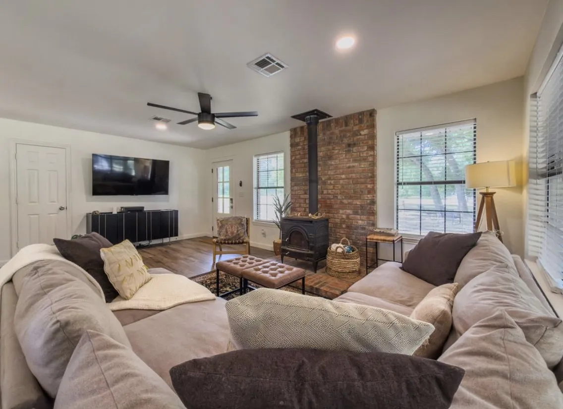 Living area with a brick accent wall, wood-finish flooring, two windows with blinds, and a ceiling fan with integrated lighting