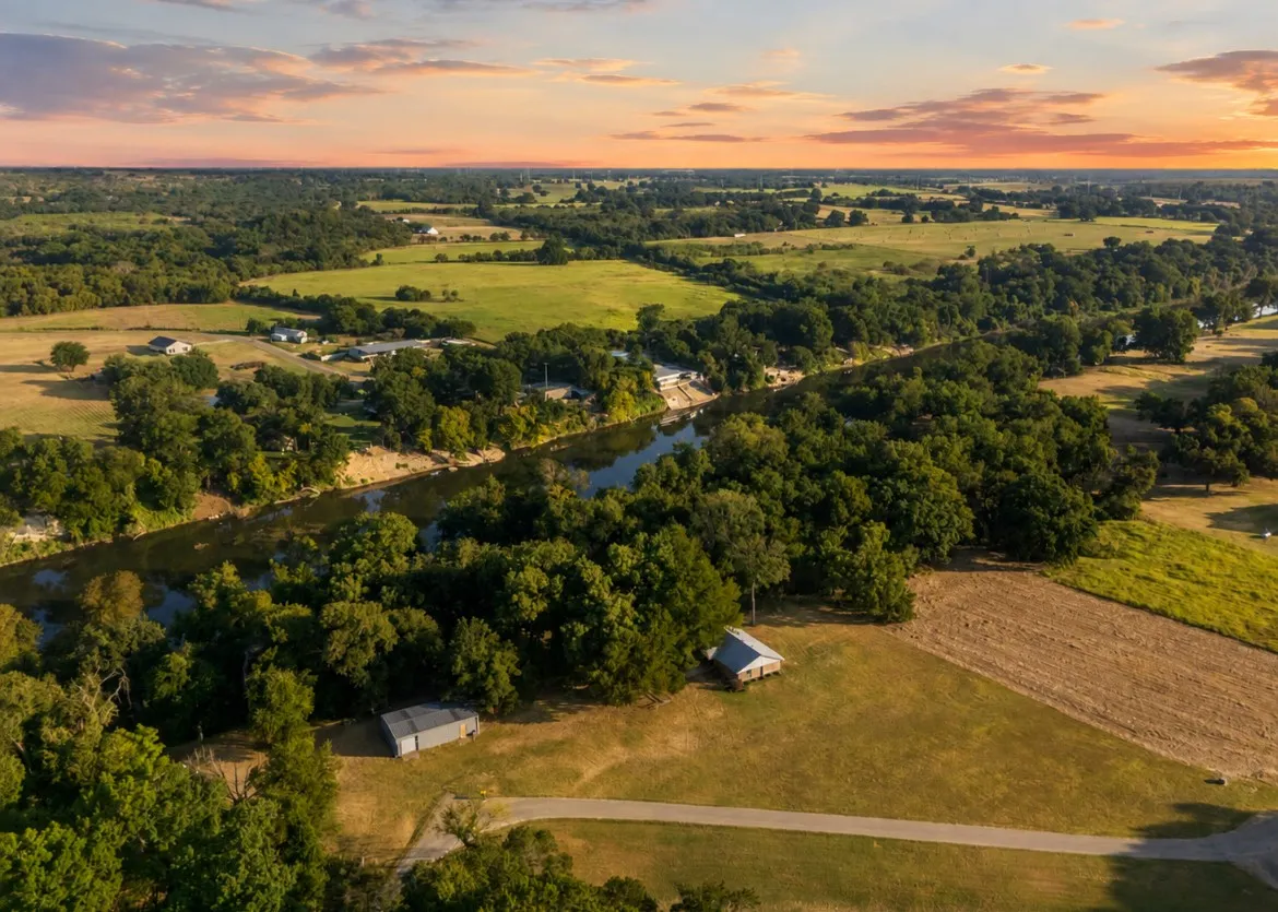 Expansive rural landscape featuring a winding river, mature trees, open fields, and scattered structures