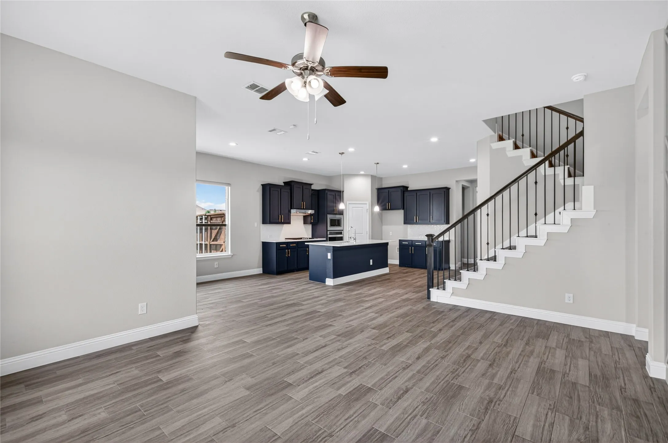 Wood-finish flooring throughout, modern ceiling fan with light fixture, recessed lighting, staircase with dark wood handrail and black balusters, and a kitchen featuring dark cabinetry and an island