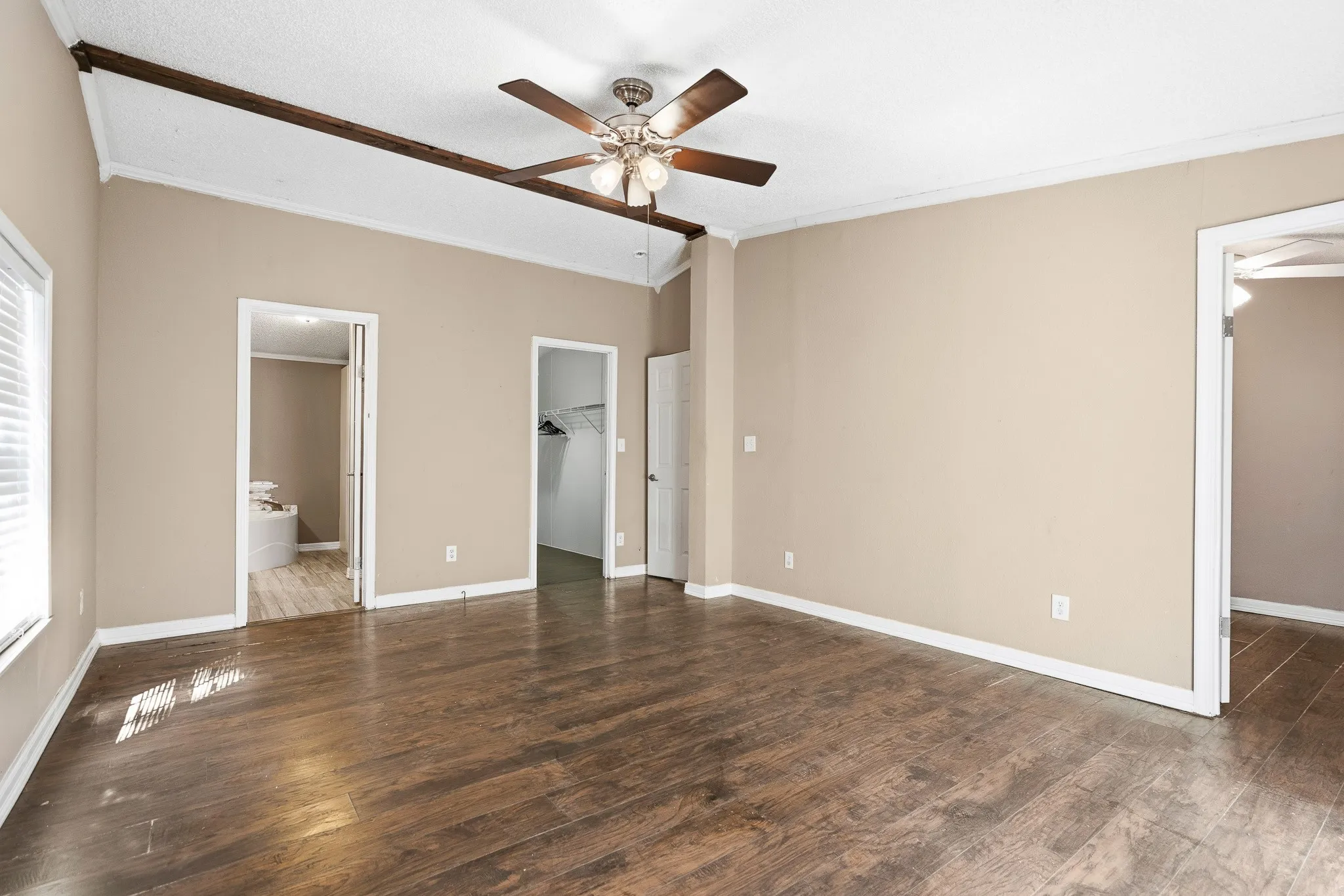 Spacious room featuring wood-finish flooring, neutral-toned walls, a ceiling fan, and exposed beam accents