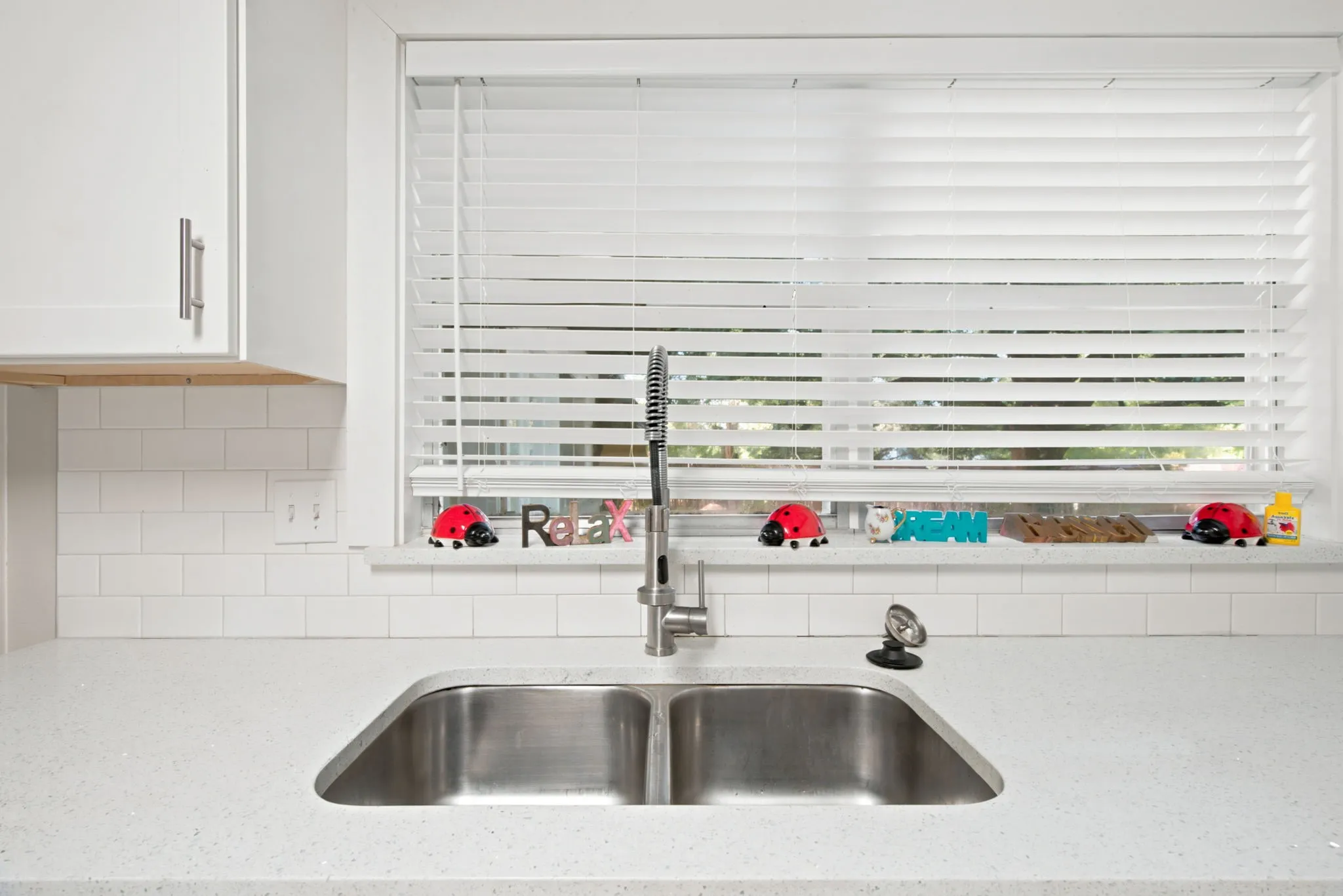 White shaker-style cabinetry, subway tile backsplash, white countertop, stainless steel double basin sink, and window with blinds