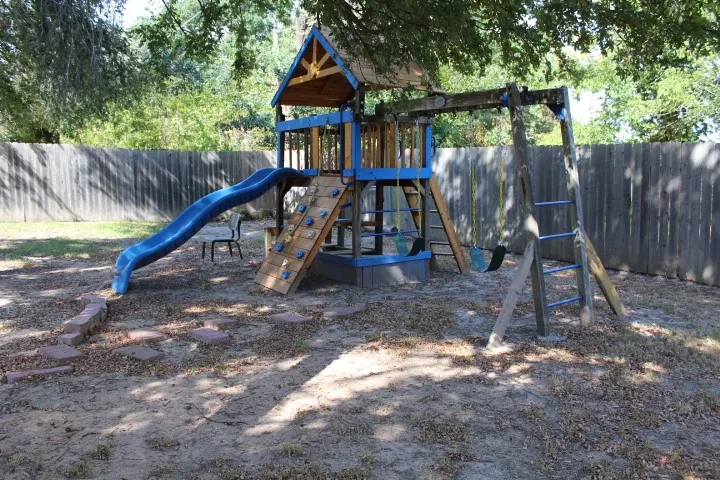 Backyard play structure featuring a slide, climbing wall, and swings, enclosed by a wood privacy fence