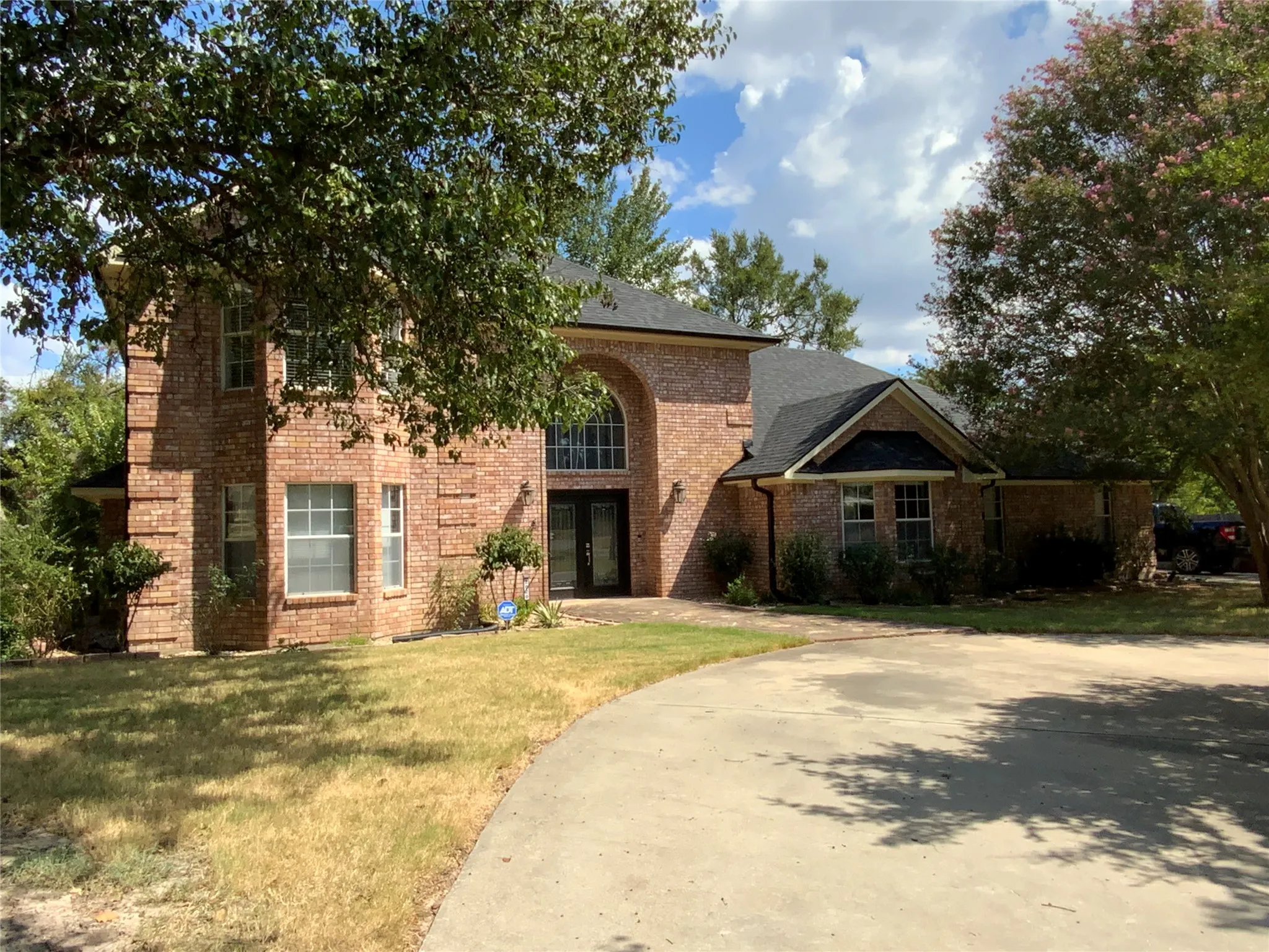 Brick facade residence featuring a prominent arched entry, dark shingled roof, and a paved driveway