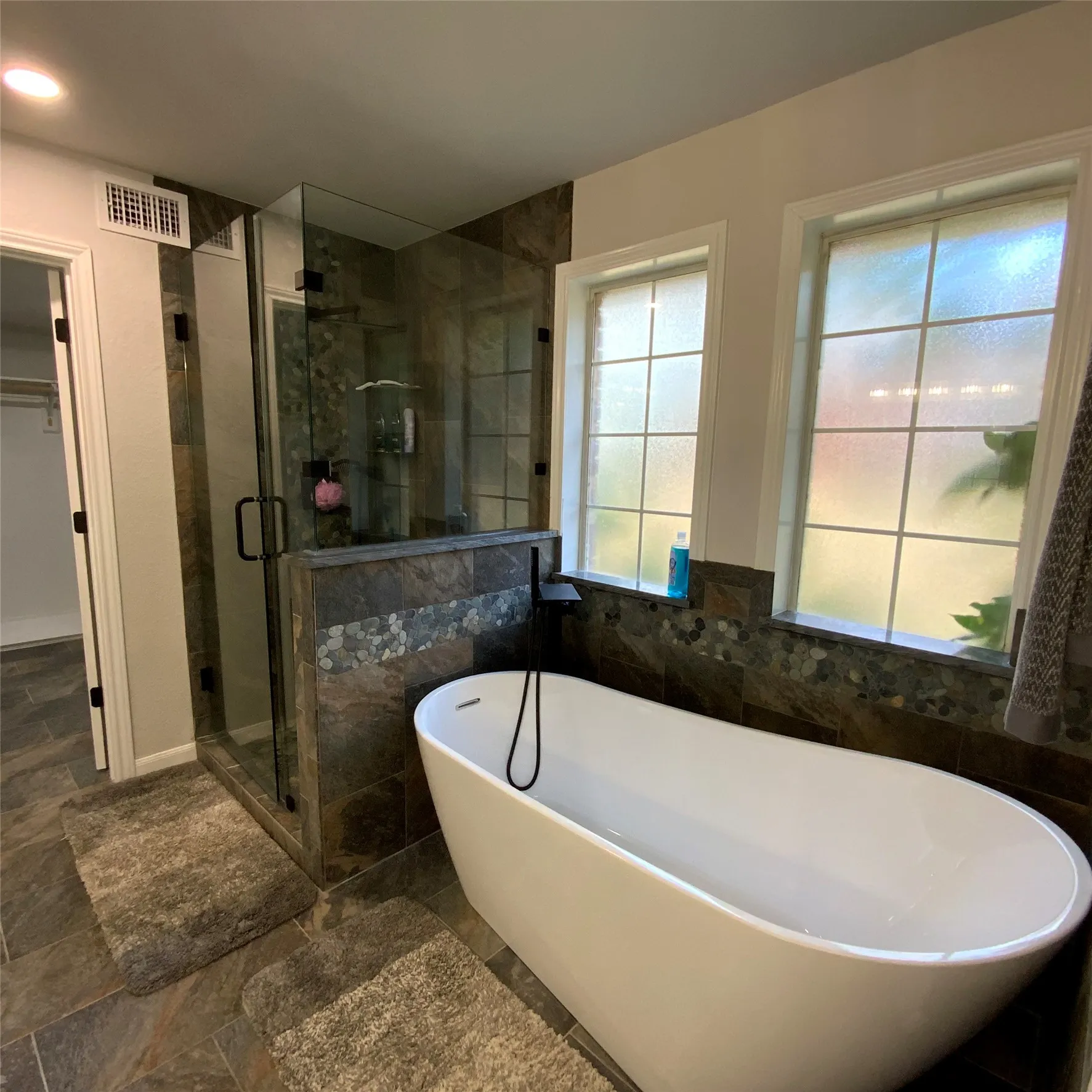 Freestanding soaking tub, floor-to-ceiling tiled shower enclosure with glass panels, dual windows, and dark-toned stone-finish flooring