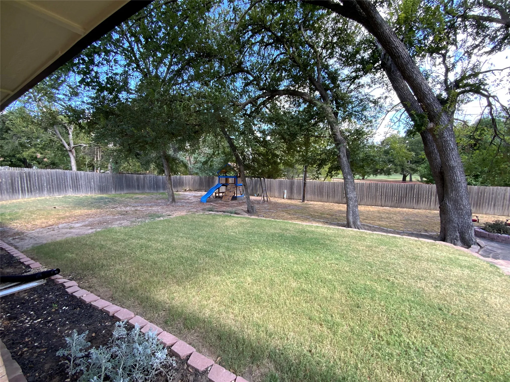 Expansive backyard featuring a privacy fence, mature trees, and a landscaped garden bed with brick edging