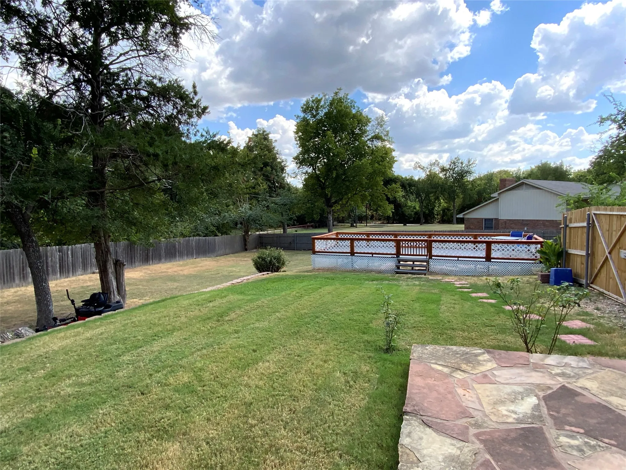 Expansive backyard featuring a manicured lawn, established trees, private wood fencing, stone patio, and a swimming pool with a surrounding deck