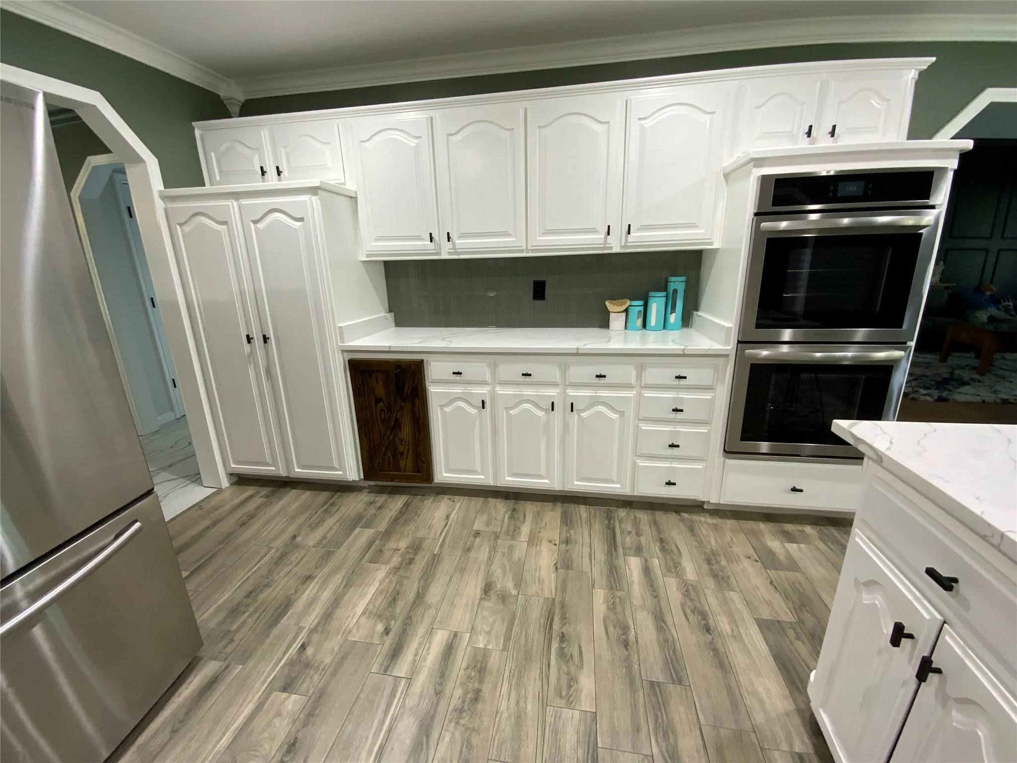 Kitchen featuring white cabinetry, wood-finish flooring, light-colored countertops, a tile backsplash, and built-in stainless steel double ovens