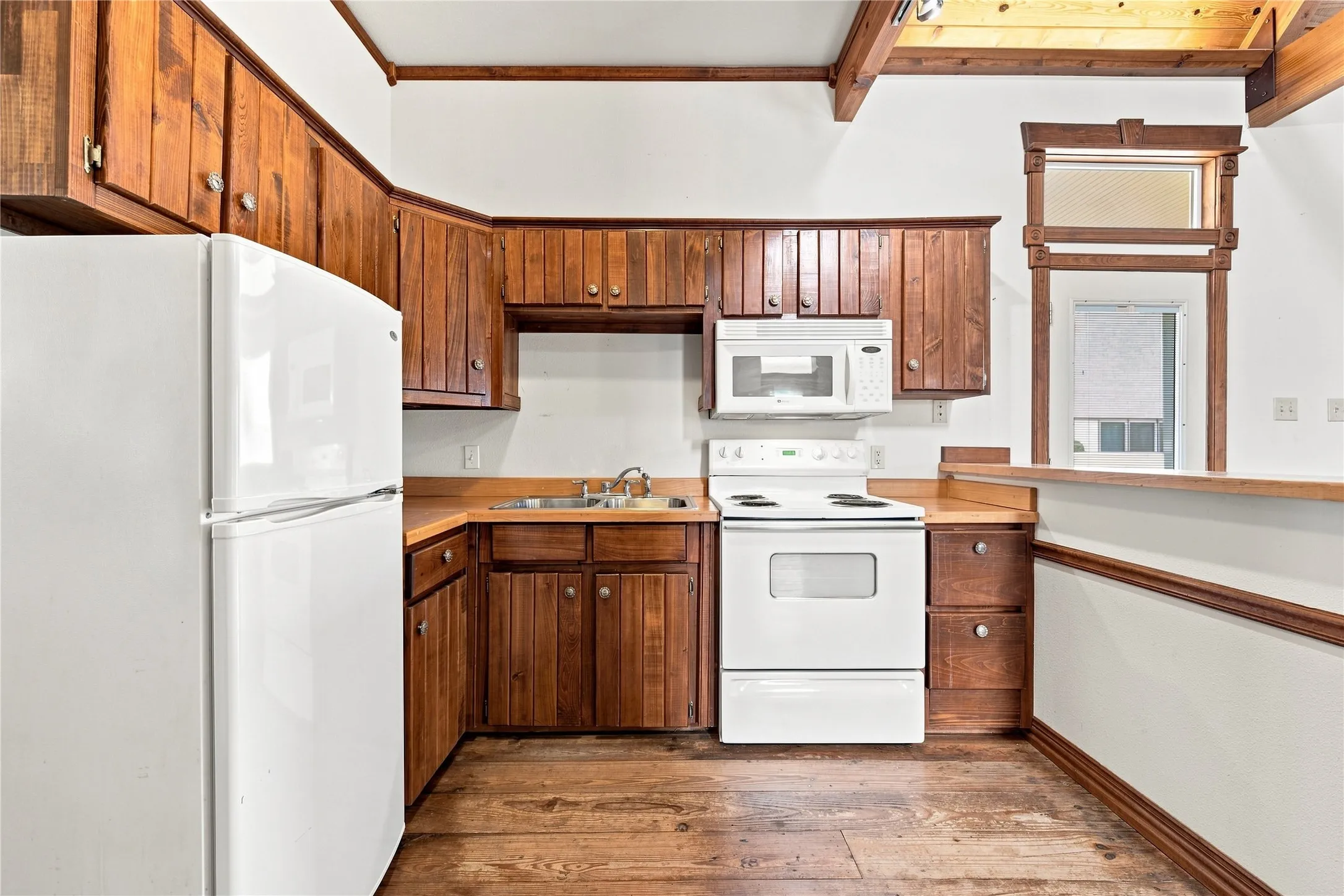 Kitchen featuring wood-finish cabinetry, white appliances, wood-finish flooring, and exposed ceiling beams
