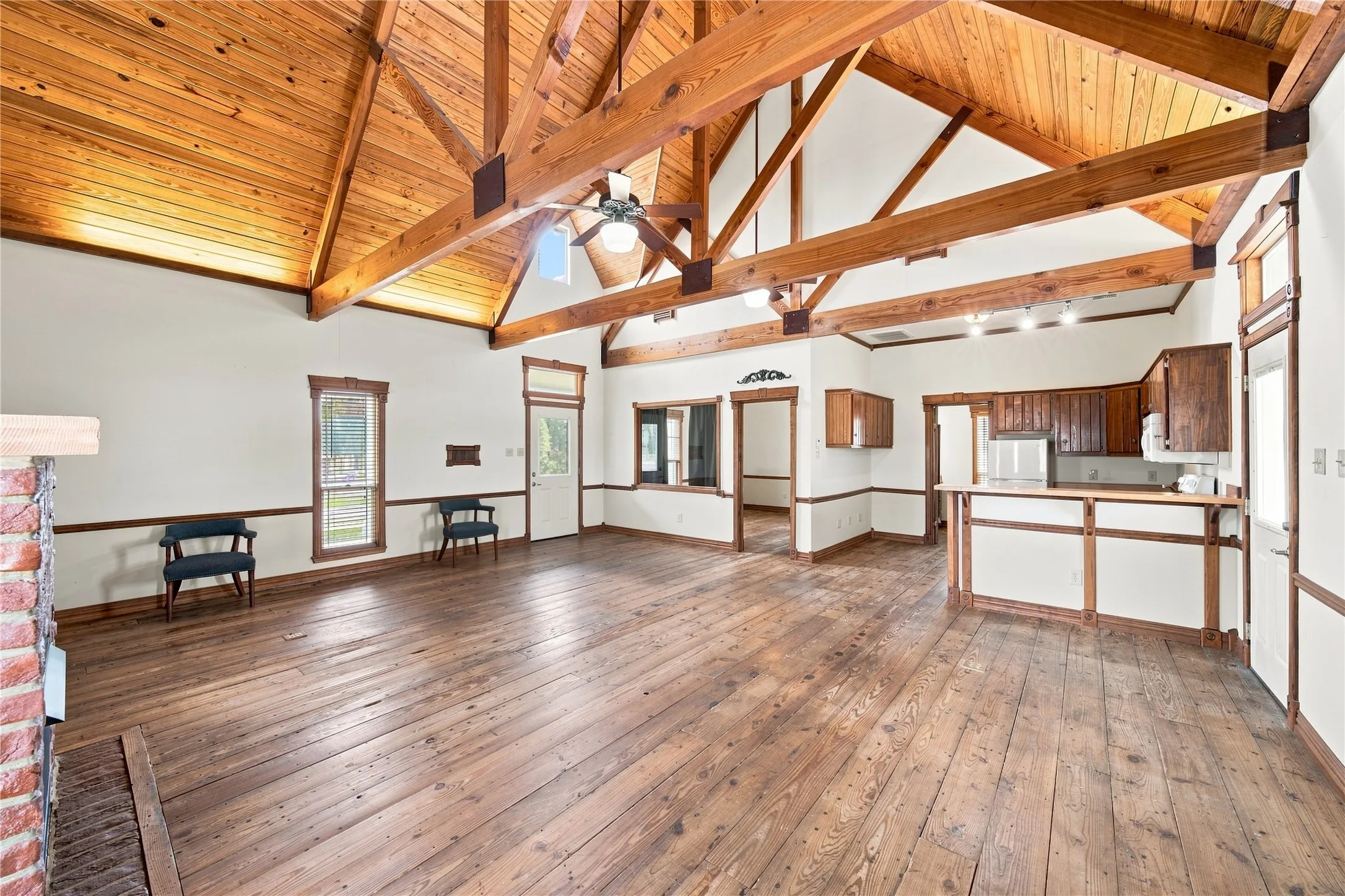 Vaulted wood-paneled ceiling, exposed wood beams, wood-finish flooring, brick fireplace, and kitchen area with wood cabinetry